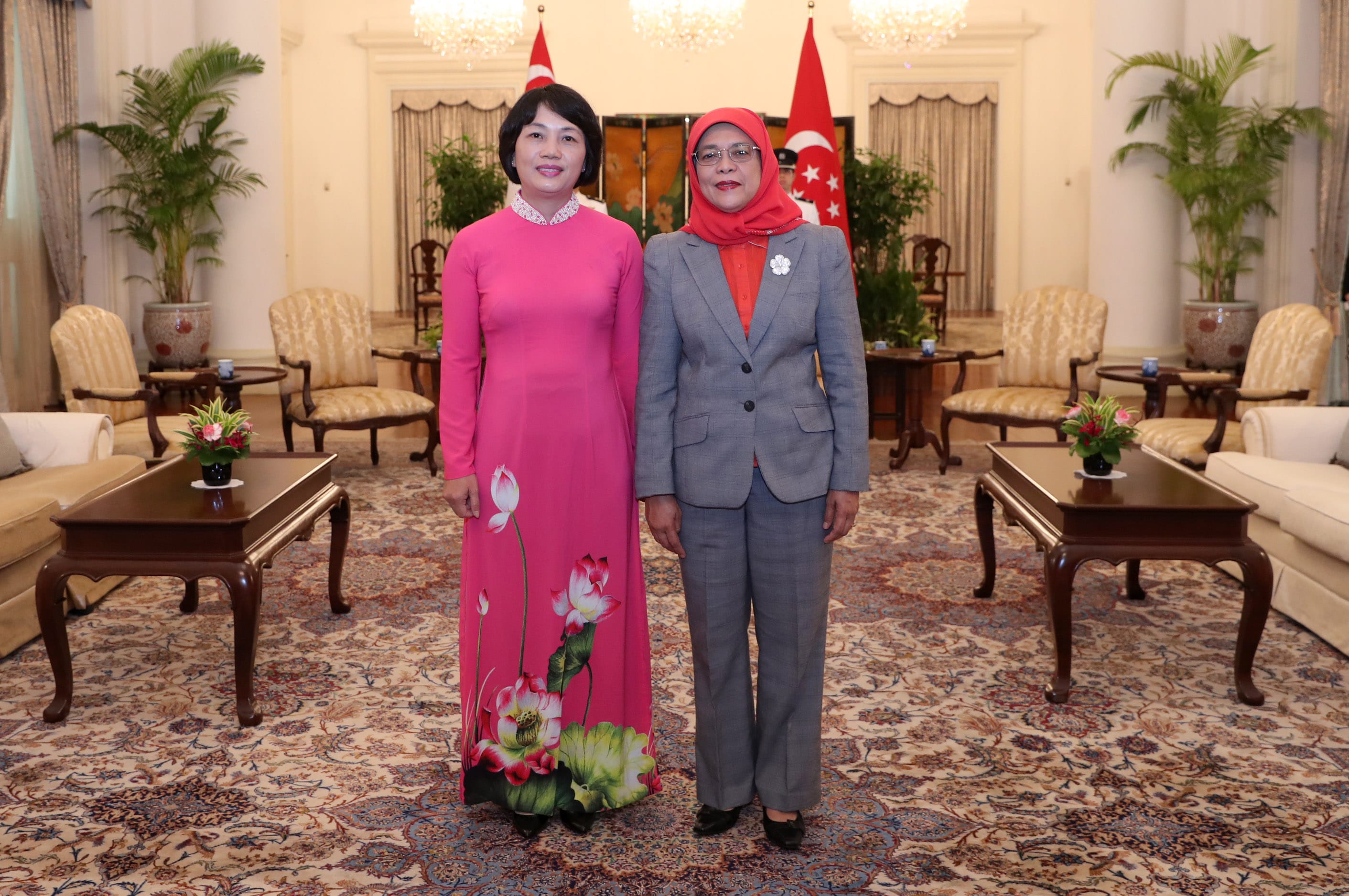 Two women standing in formal attire, one in a pink dress with floral details, the other in a gray suit and red hijab.