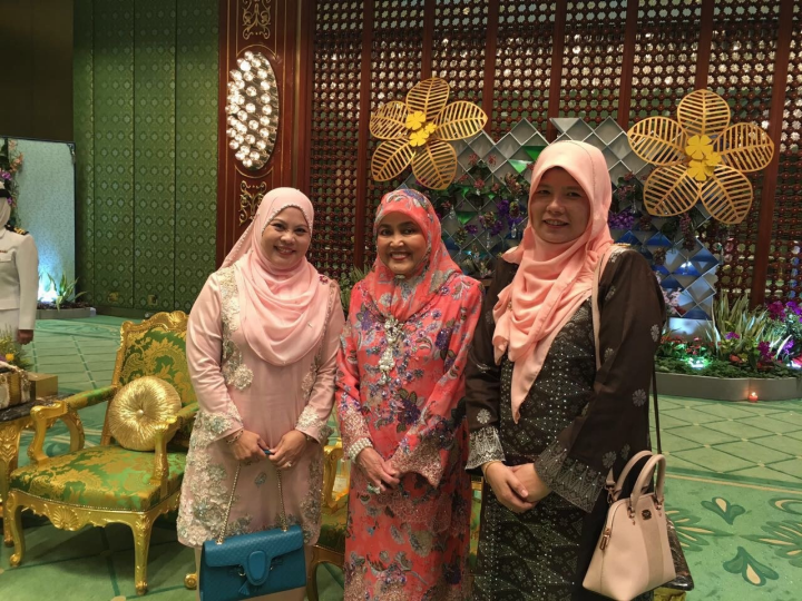 Three women in headscarves pose in front of a decorative wall; a man in uniform stands to the side.