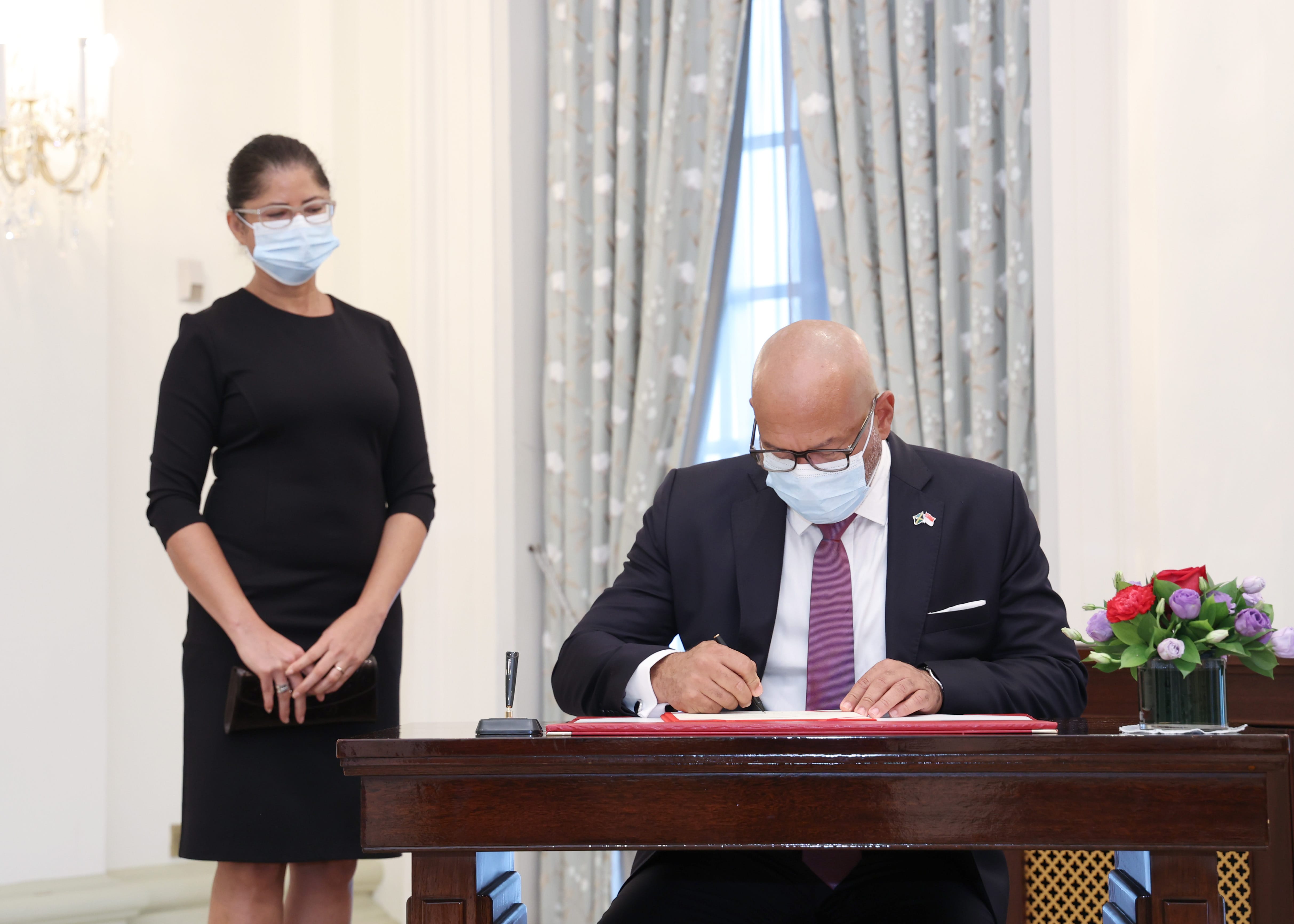 President Solih signs document at desk, wearing a mask and suit. A woman in a black dress stands nearby.