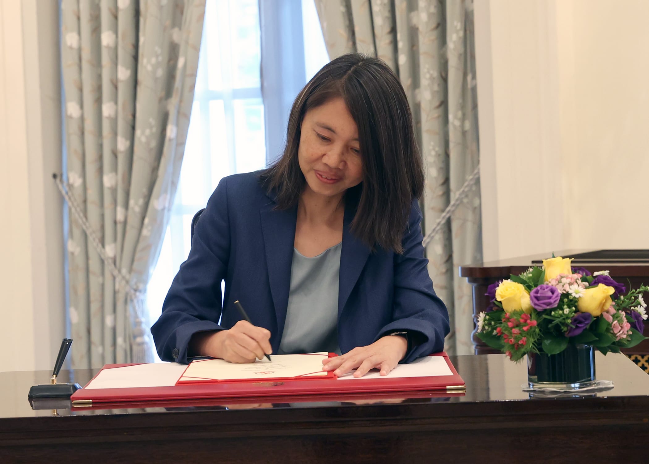 Woman in a blue blazer signing a document on a table with flower arrangement.