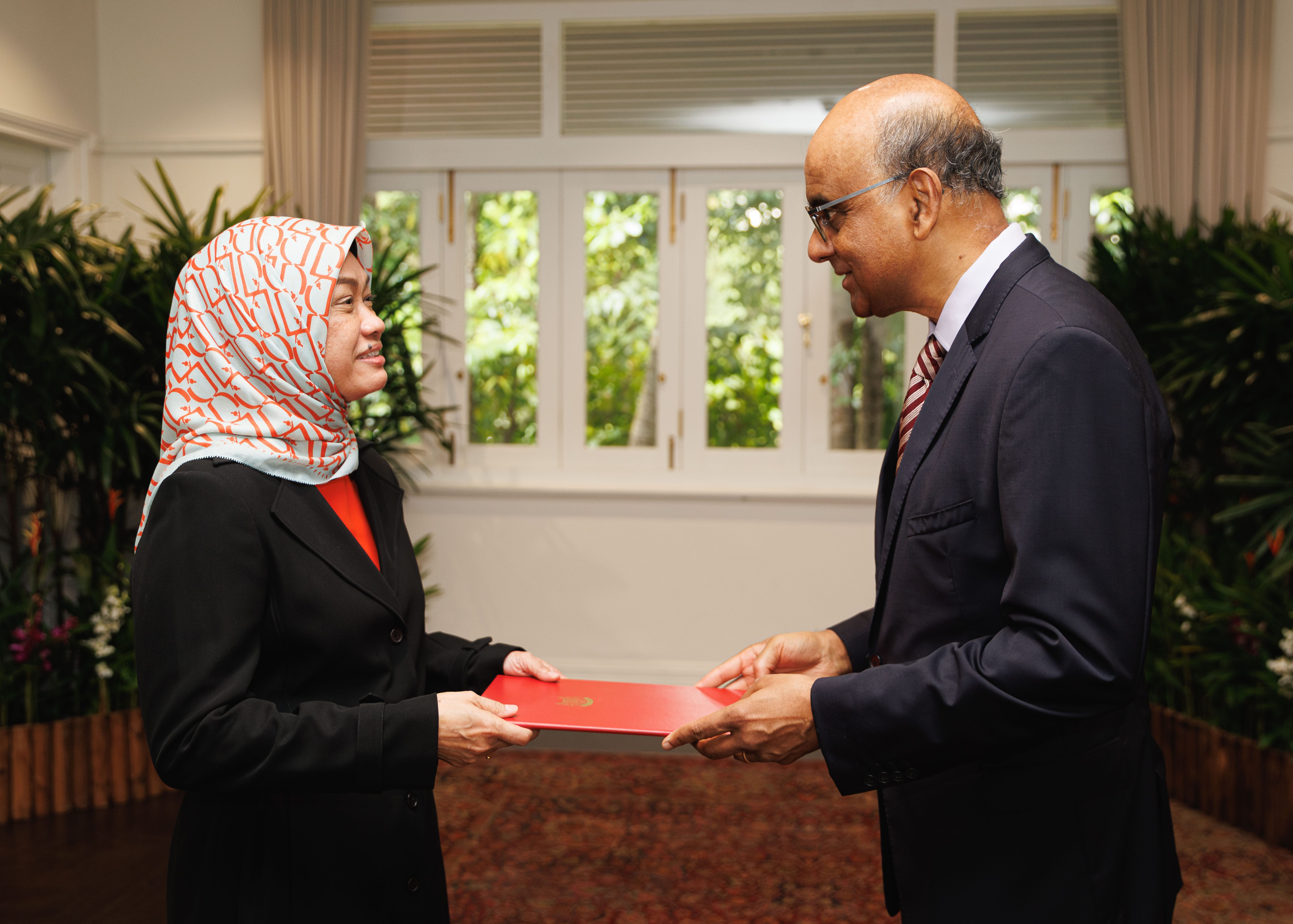 Two formally dressed people exchanging a red document. Woman wears a patterned hijab.