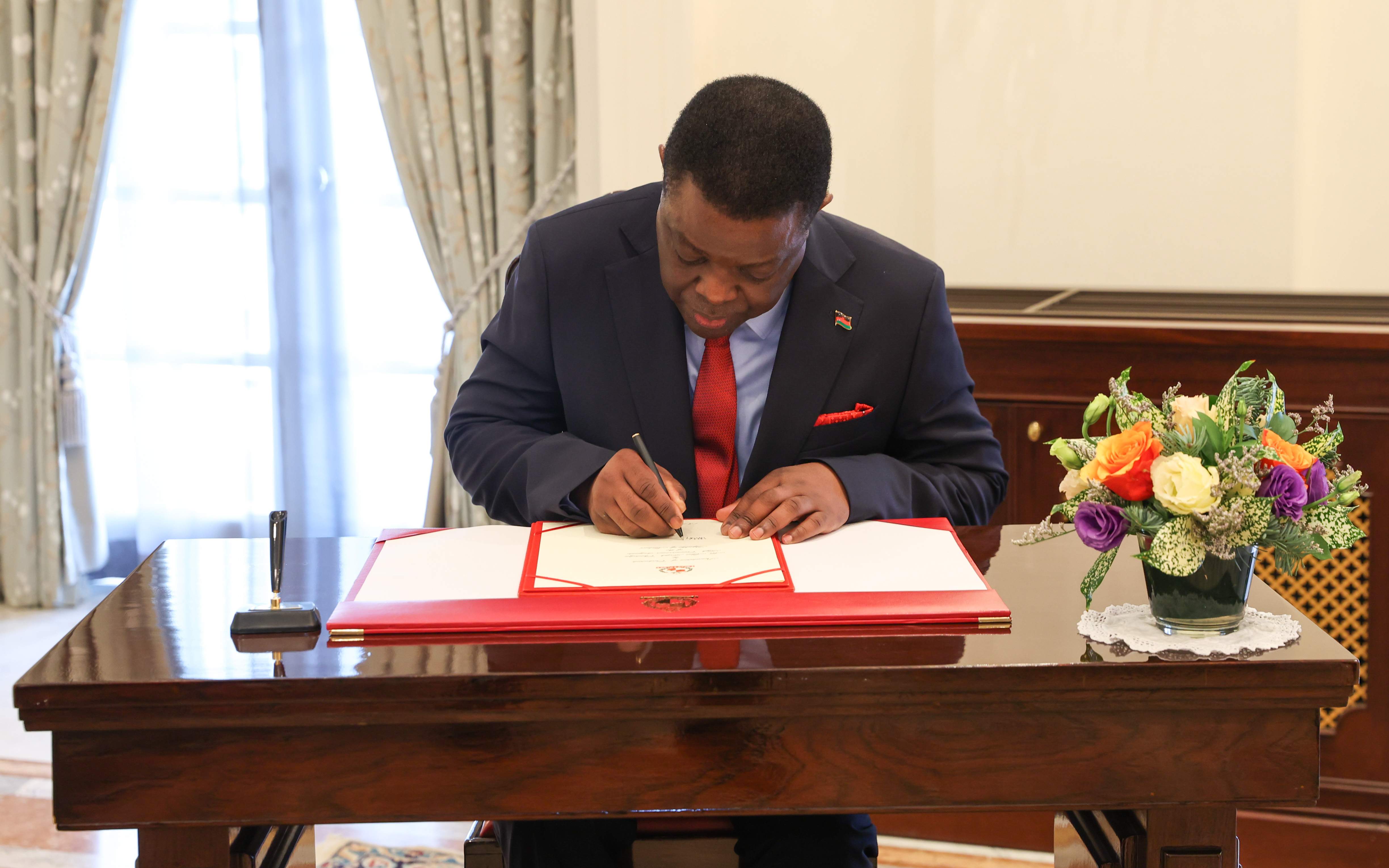 Lazarus Chakwera signing a document on a red pad, wearing suit with a Malawi flag pin.