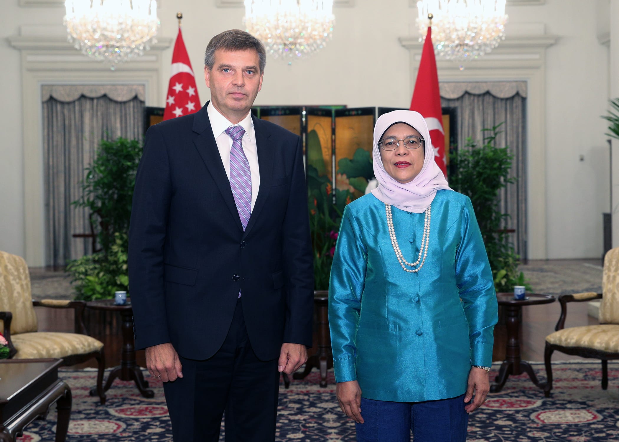 Man in suit, woman in headscarf and pearls pose indoors with Singapore flags.