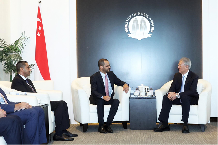 Men in suits sit in white chairs, facing each other. Singapore flag and Ministry of Home Affairs logo are visible.