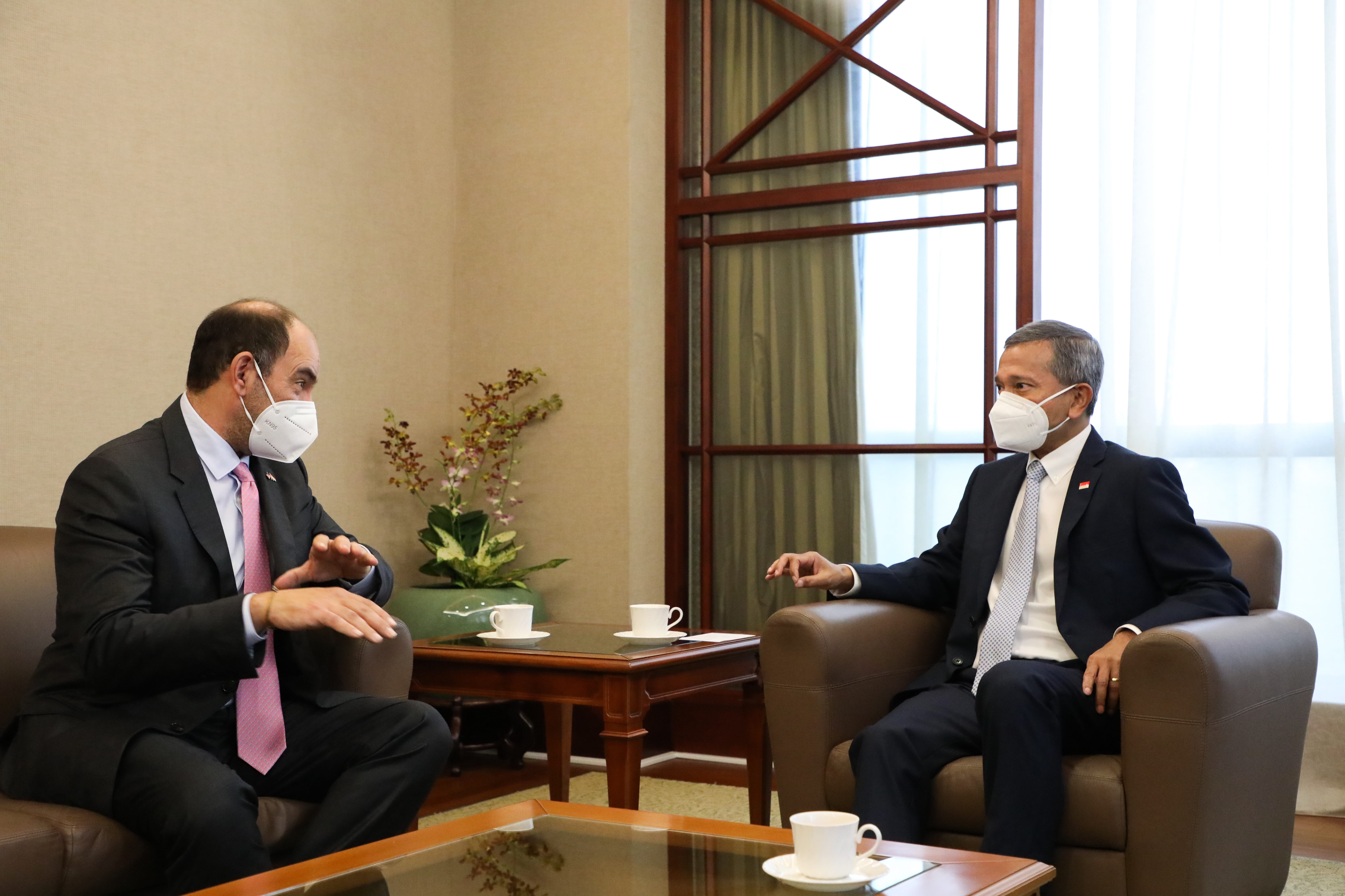 Two masked men in suits sit in armchairs, conversing over coffee on a table.