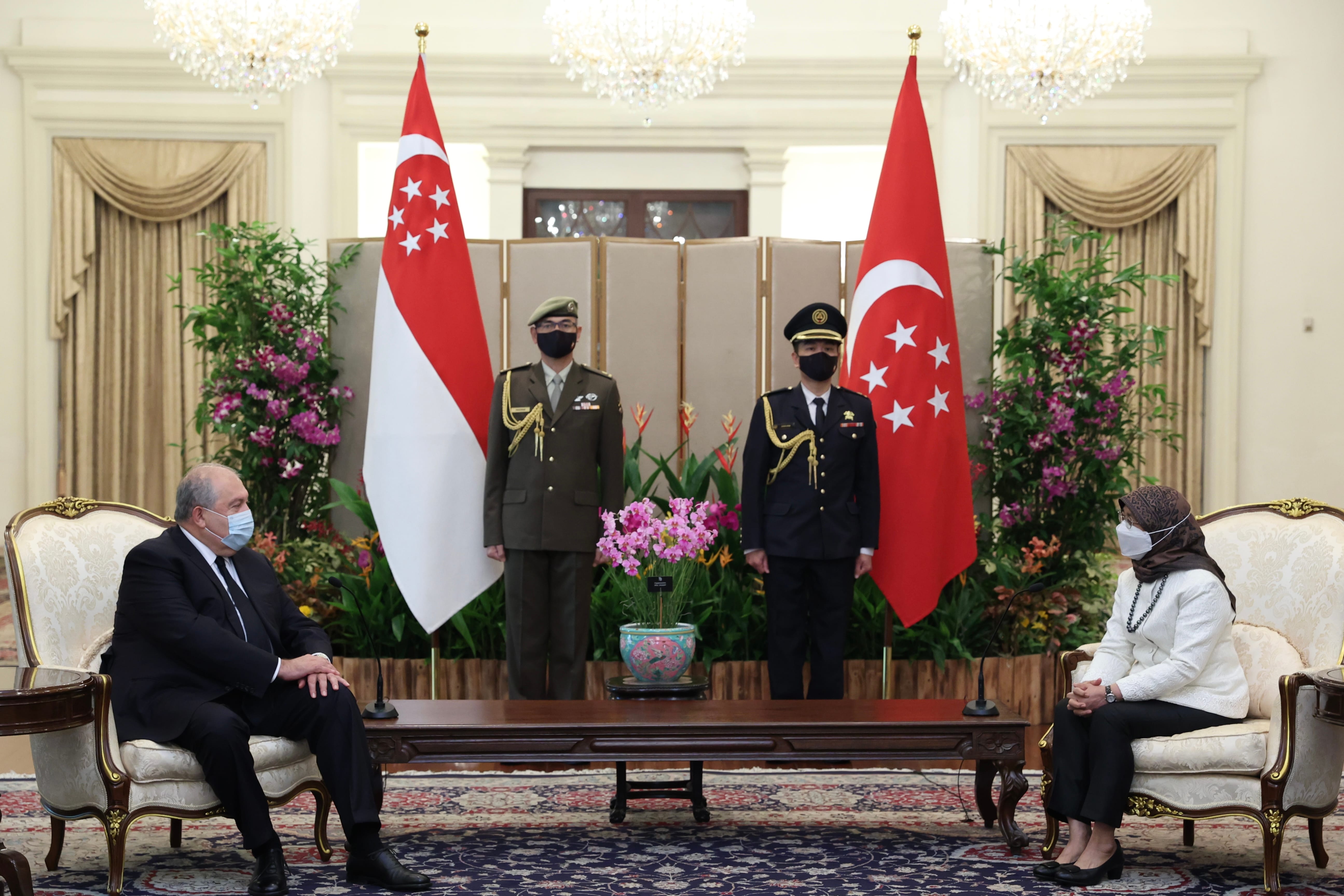Two people in suits and two in uniform sit under Singaporean flags in an ornate room.