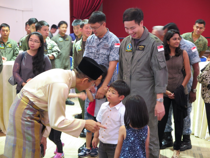 Man in traditional attire interacts with children as uniformed military personnel stand by.