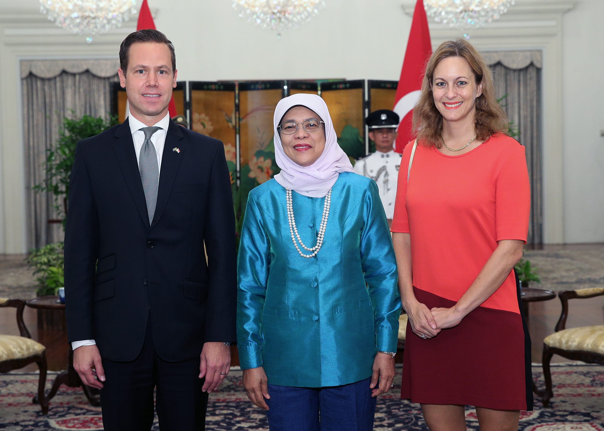 Three people stand indoors with flags and a guard in the background.