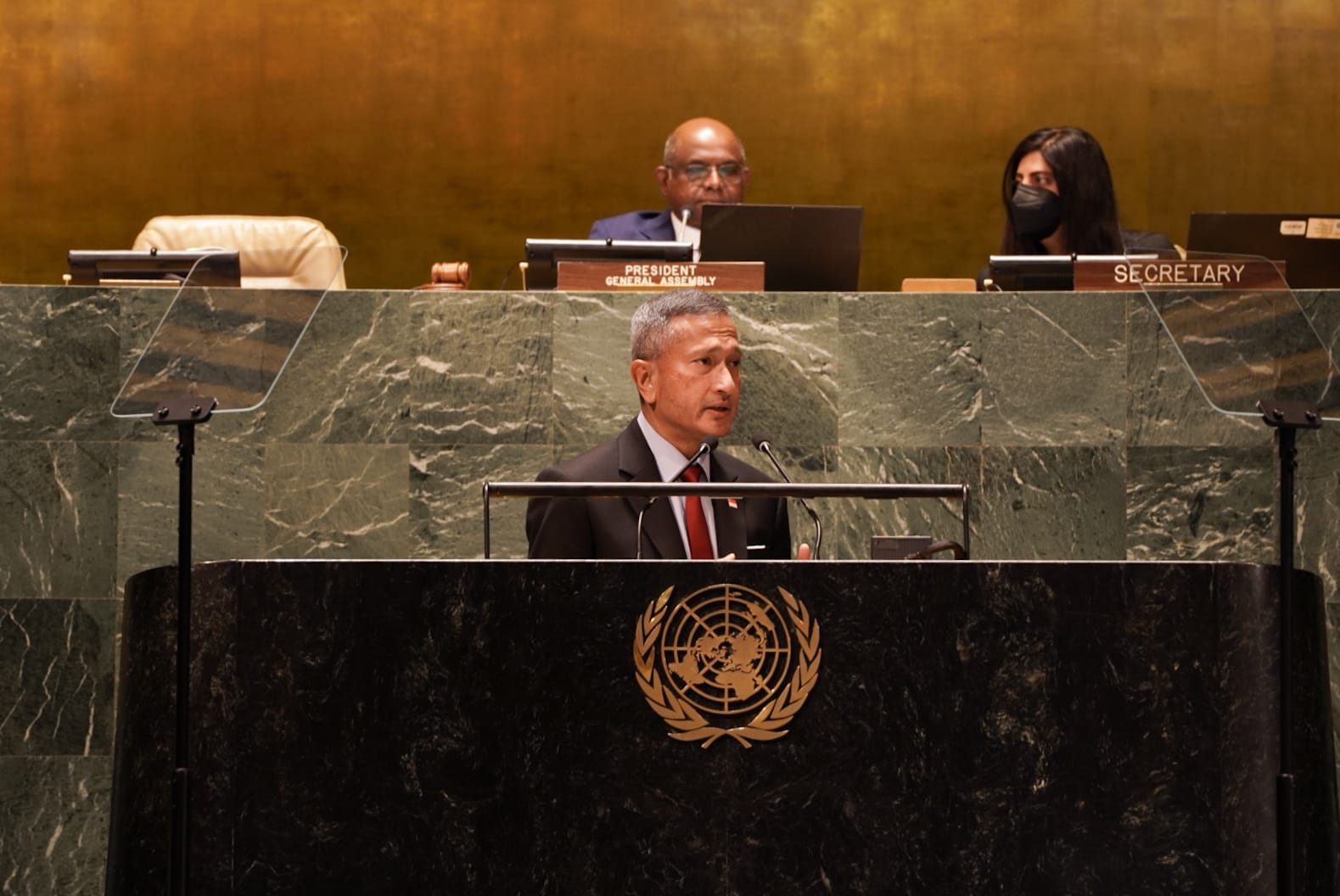 Man at UN podium with logo speaks; President General Assembly & Secretary sit behind on upper tier.