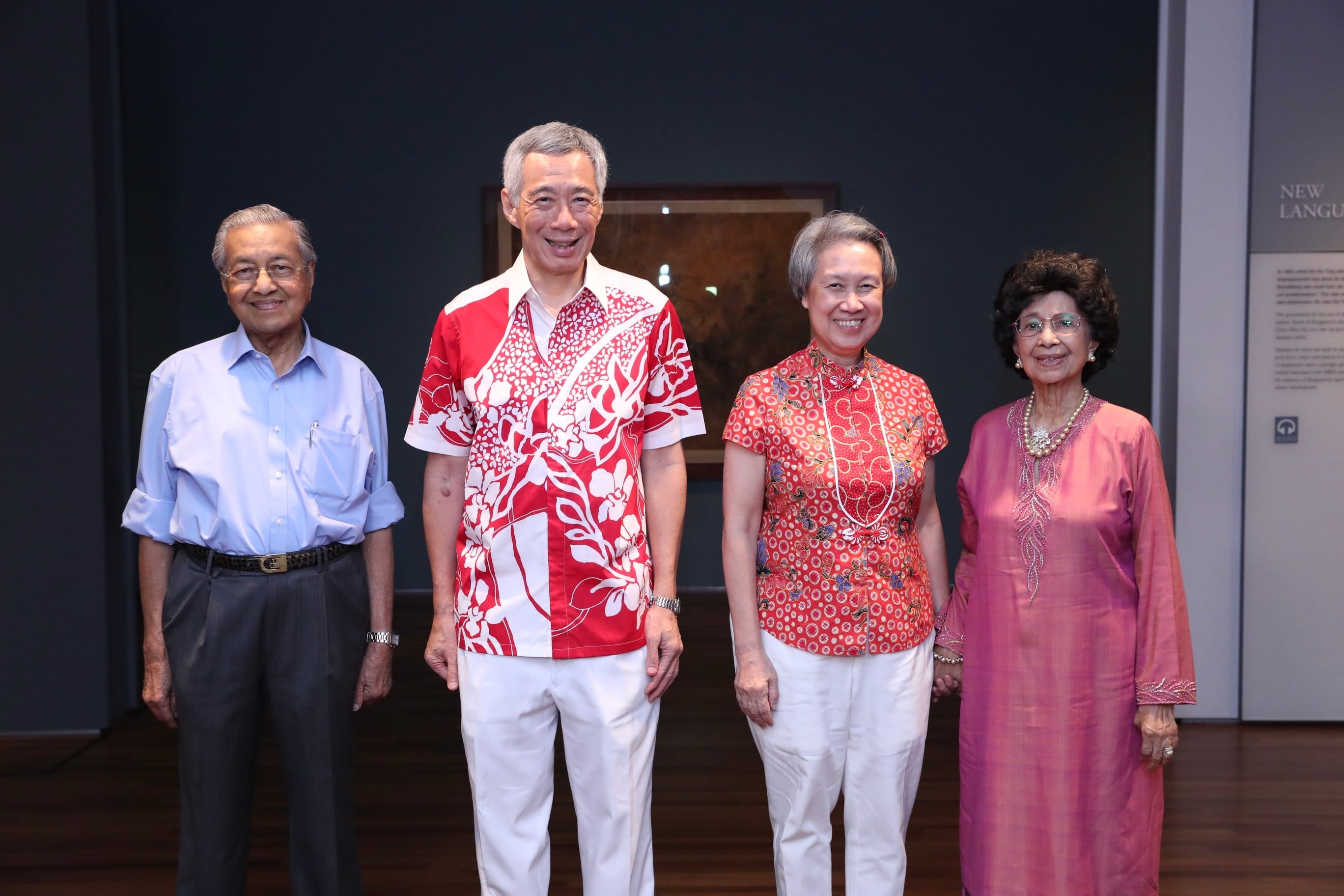 Four older adults stand side-by-side. Two men, two women, with red patterned shirts on the two figures in the middle.