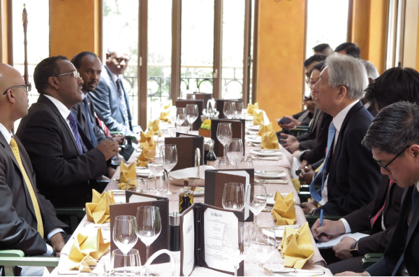 Business meeting at long set table. Two groups in suits face each other.