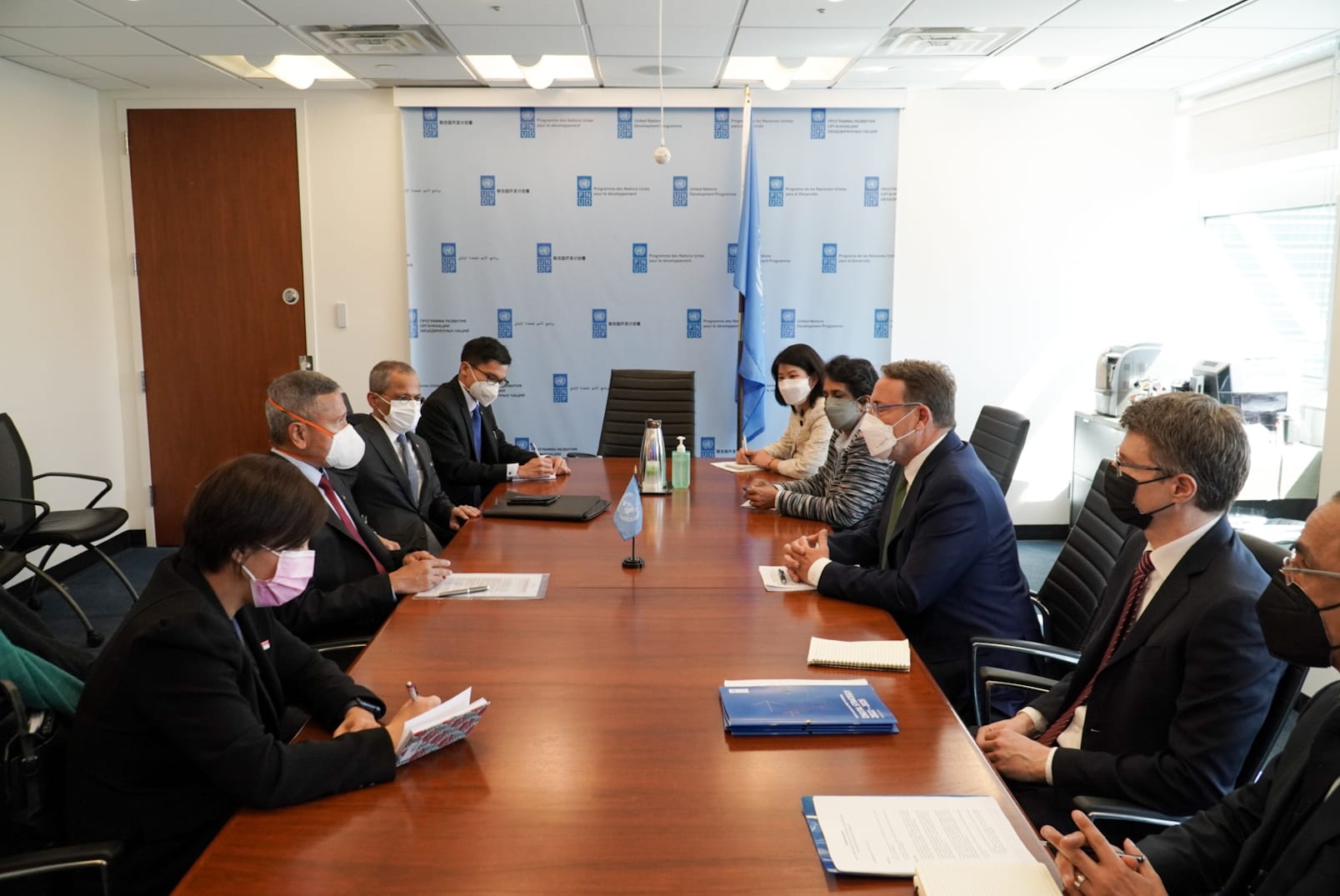 People in masks at a wood conference table with UN logos in background.