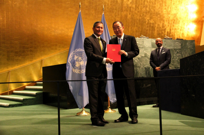 Ban Ki-moon receives document with UN flags behind them in a golden room.