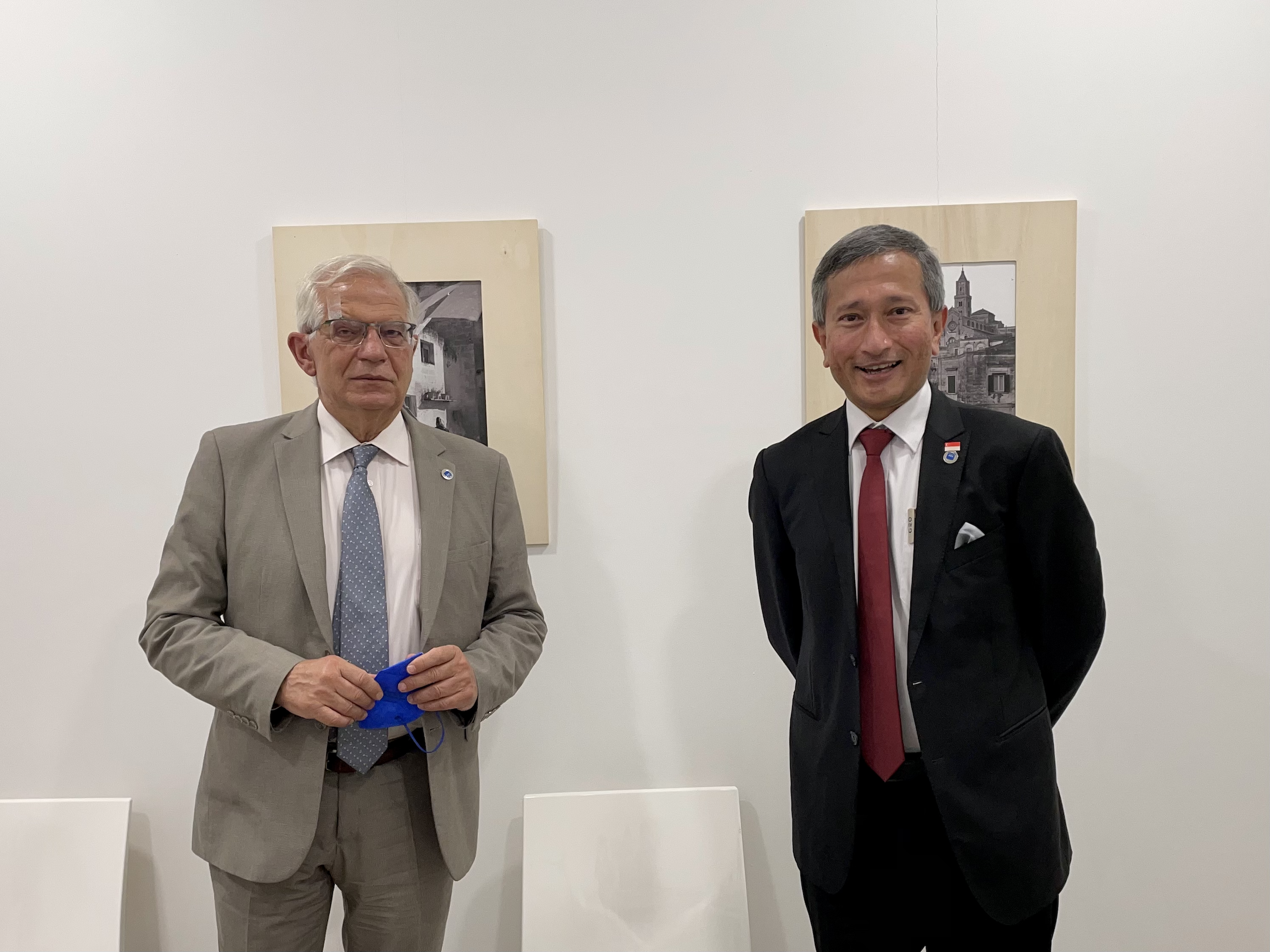 Two men in suits pose in front of framed art; one holds a blue mask.