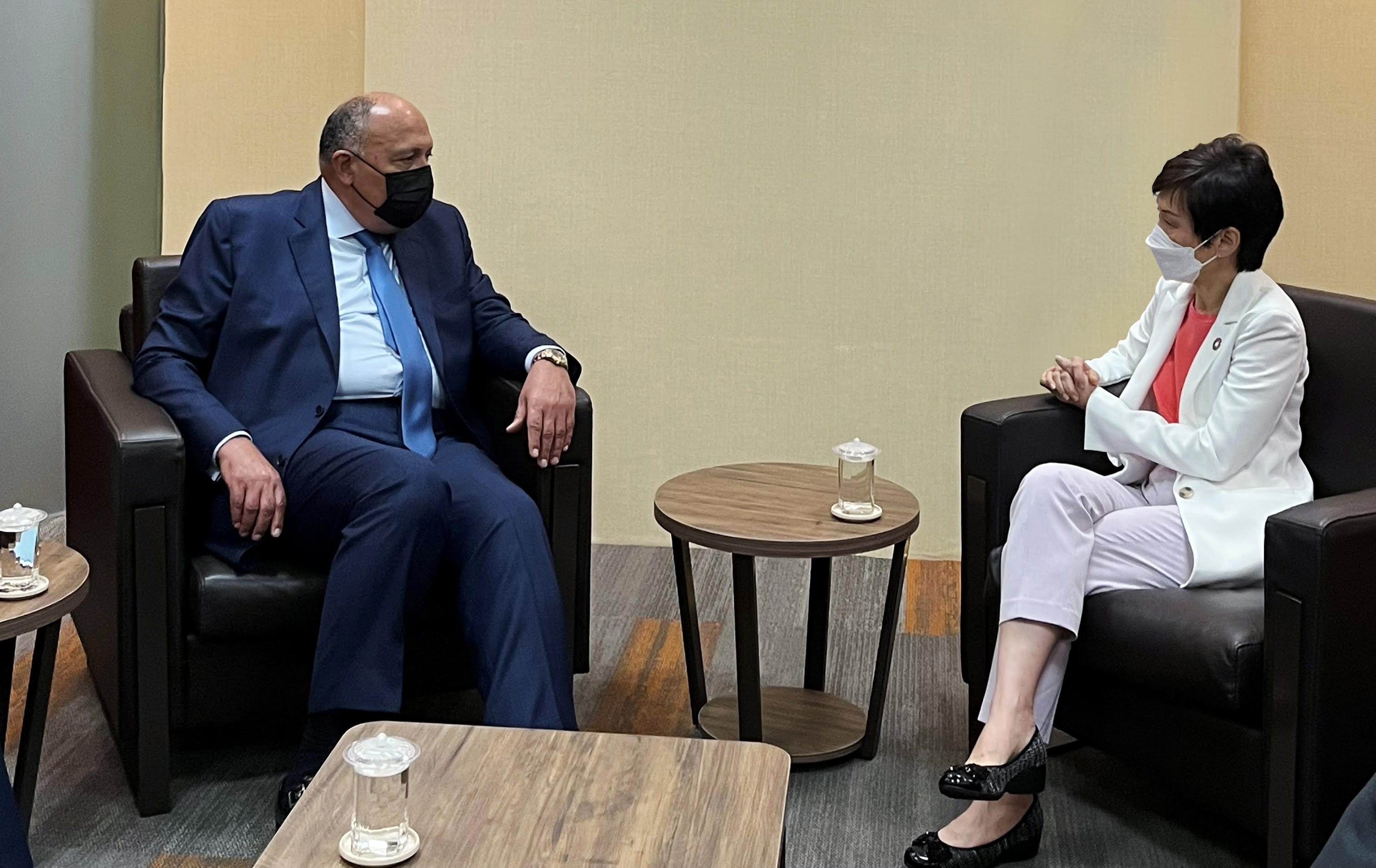 Two masked people sit in chairs facing each other, separated by a small round table.