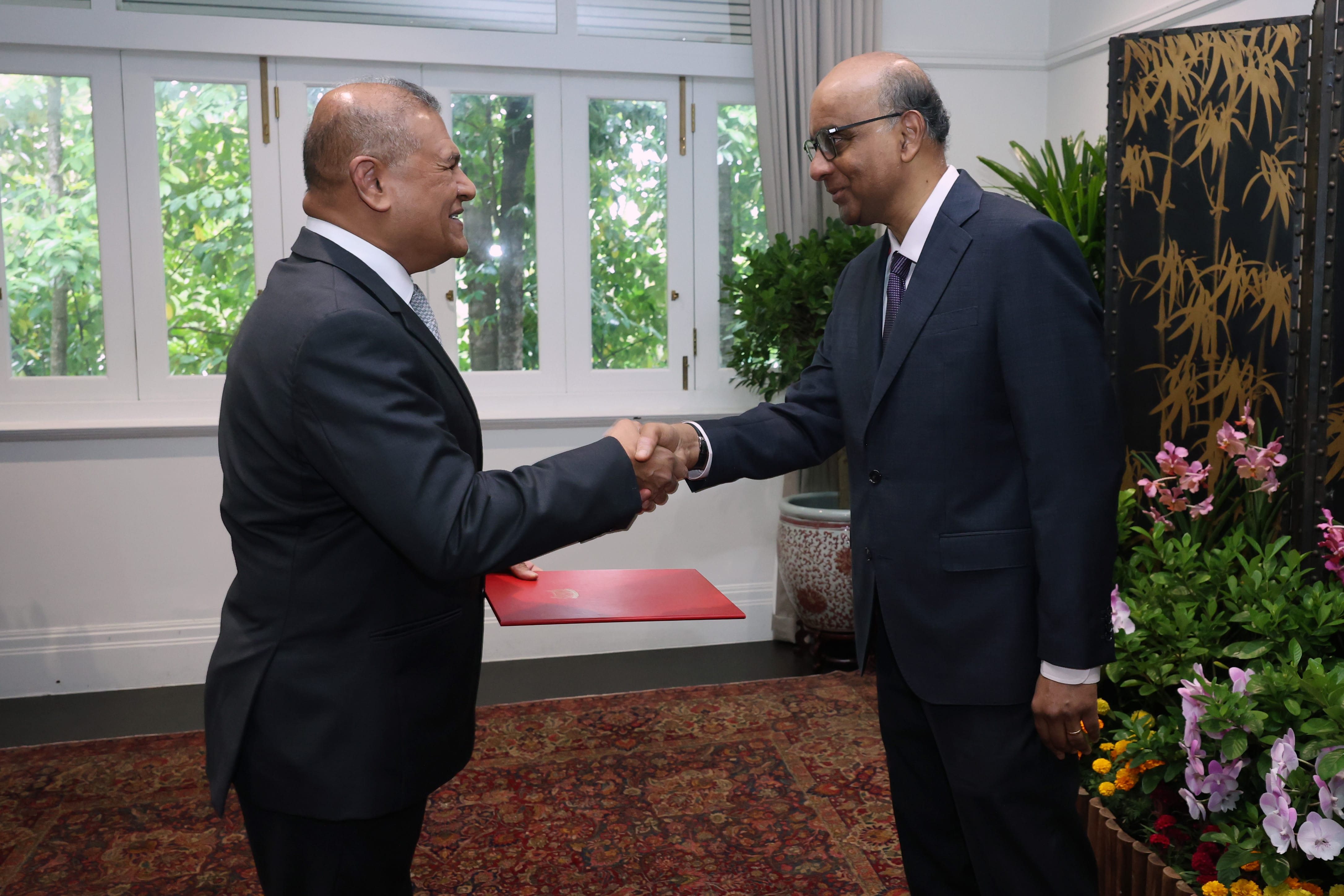Two men in suits shake hands, one holding a red folder. Window, plants, and screen in background.