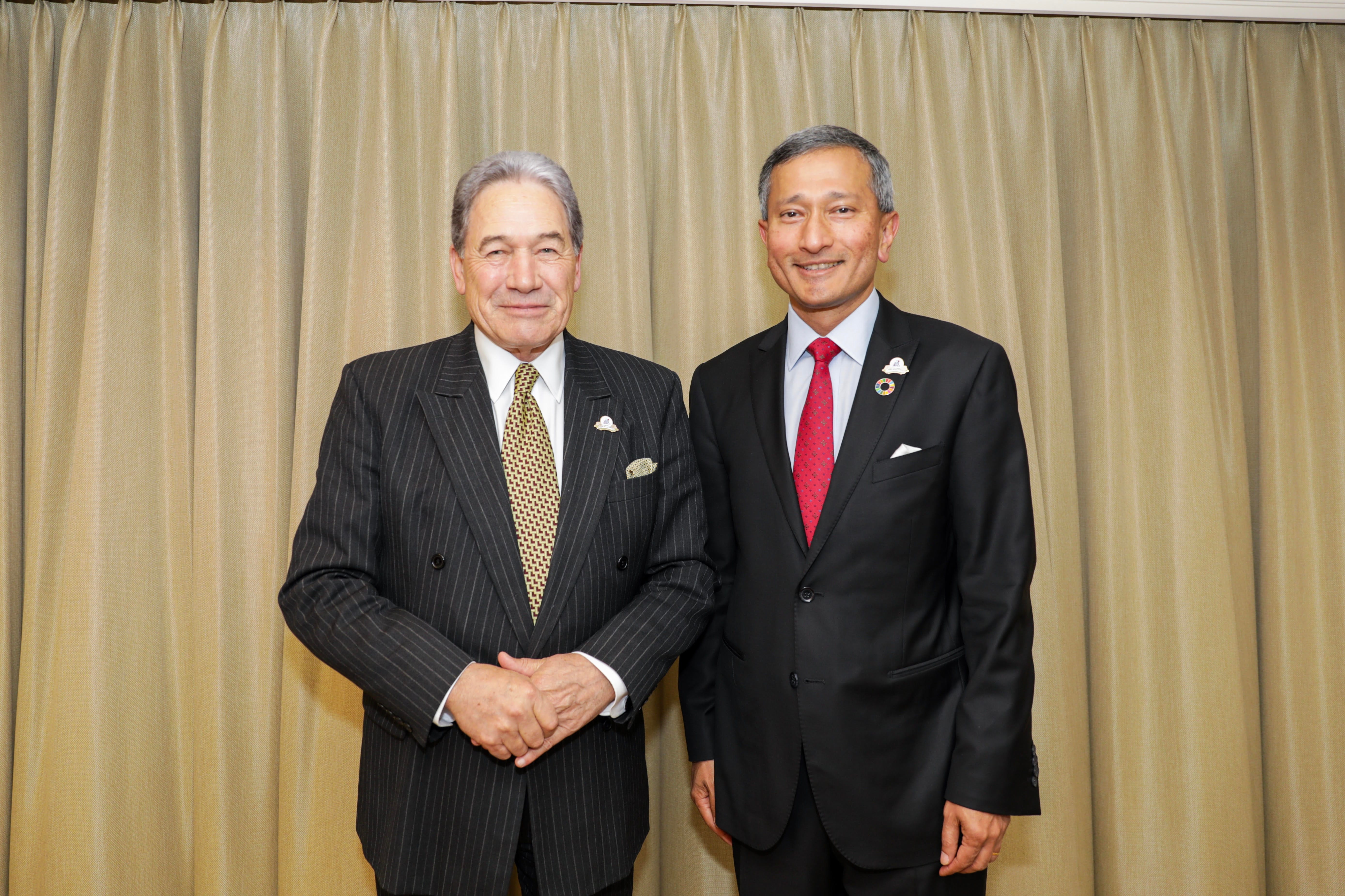 Two men in suits stand side-by-side against gold curtains. One wears a pinstripe suit, the other a solid black suit.