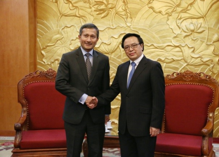 Two men in suits shake hands in front of a gold lotus wall hanging.