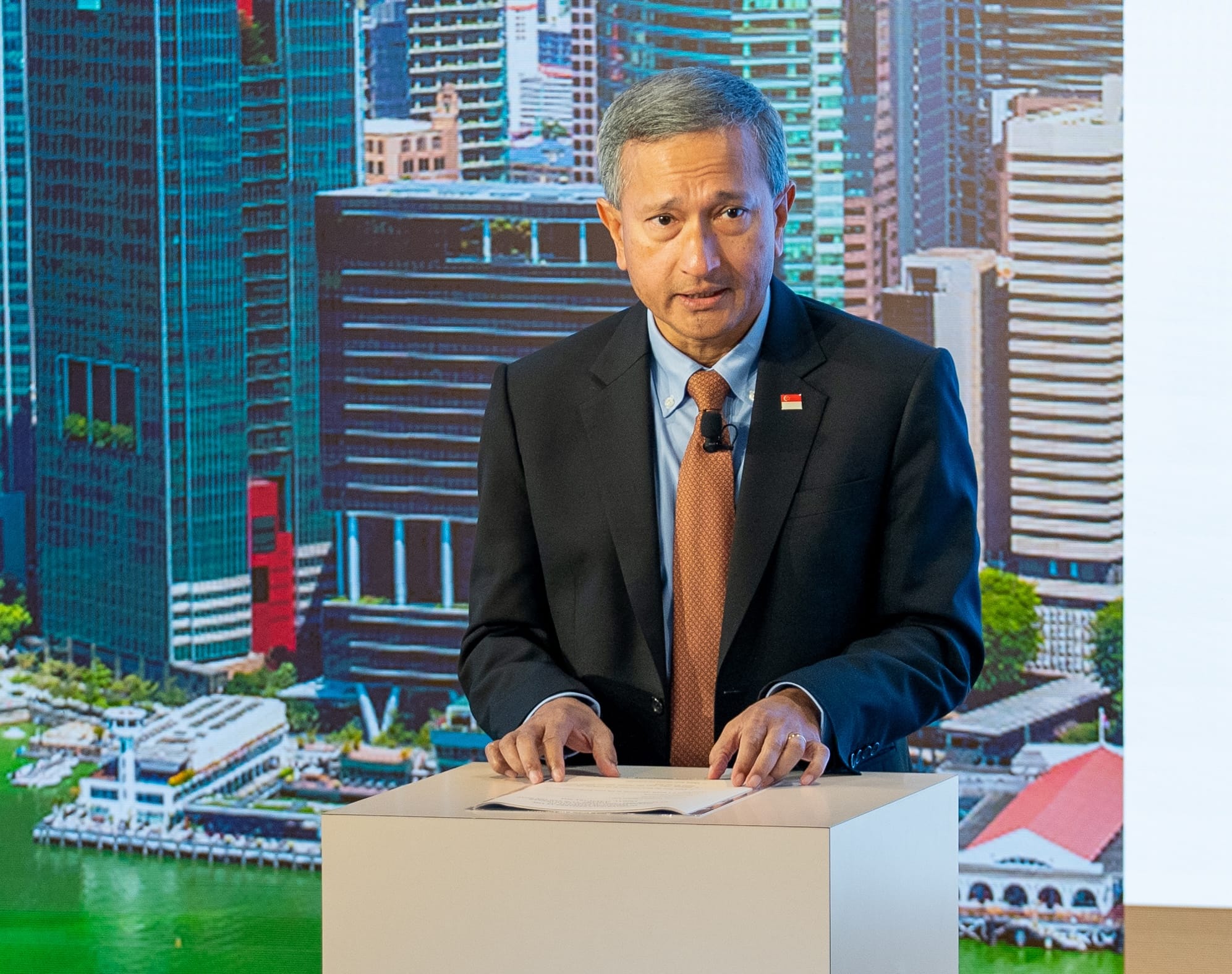 Singaporean speaker at lectern with Singapore flag pin. City skyline backdrop.