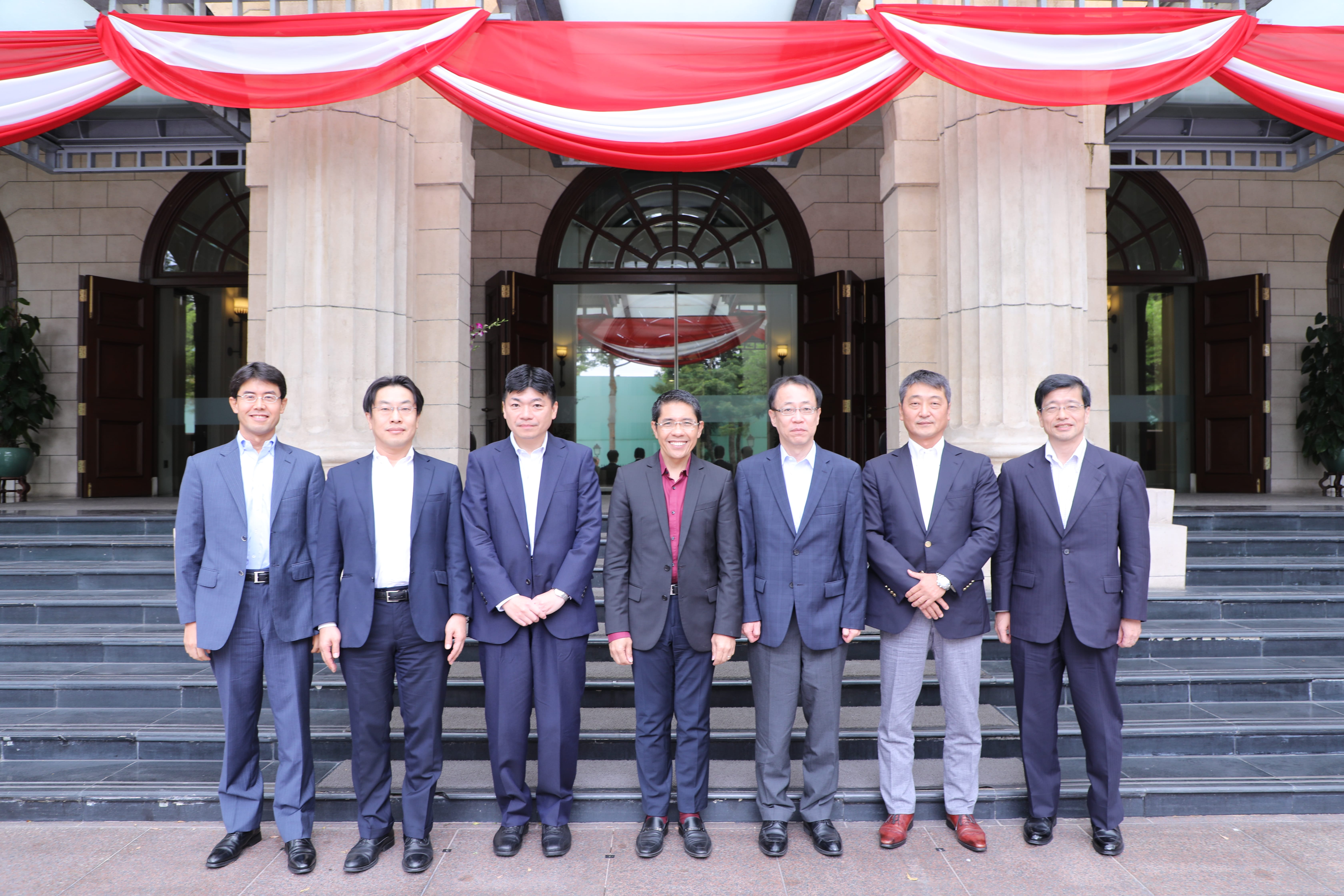 Seven men in suits stand on steps before a building decorated with red and white draped fabric.