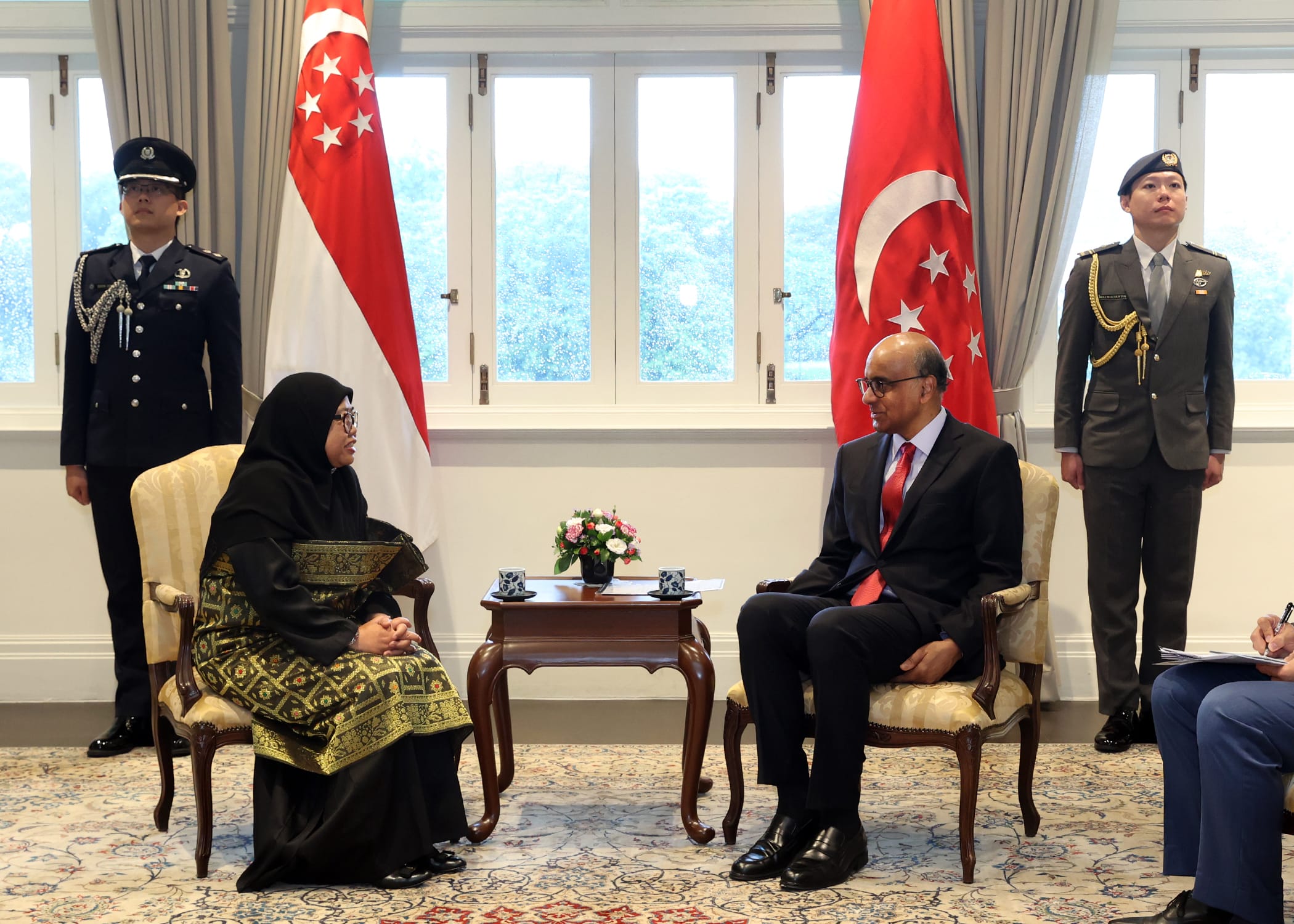 Two people seated, facing each other, flanked by Singapore flags and uniformed guards.