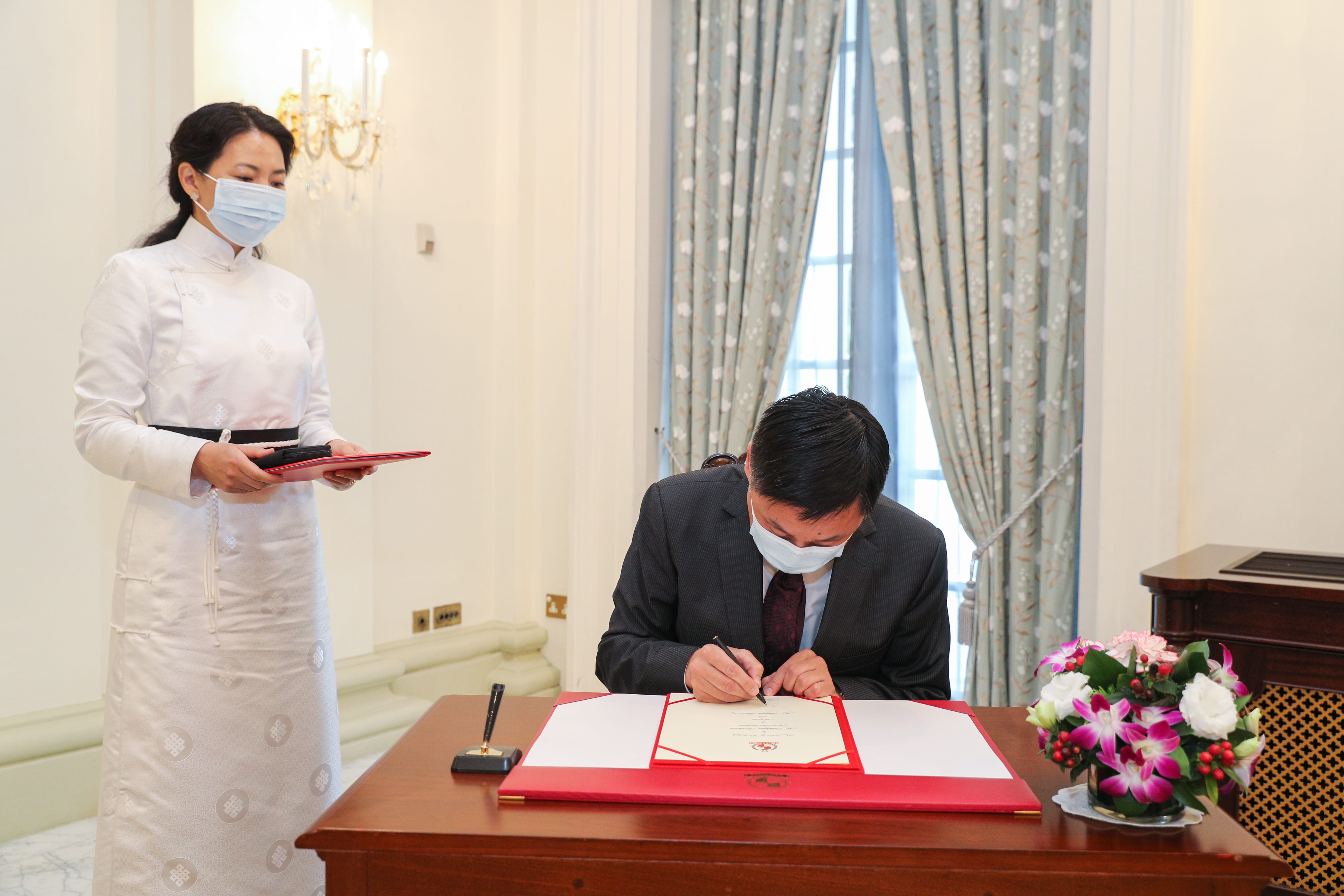 Person signing document at table, another person in white dress & mask standing nearby.