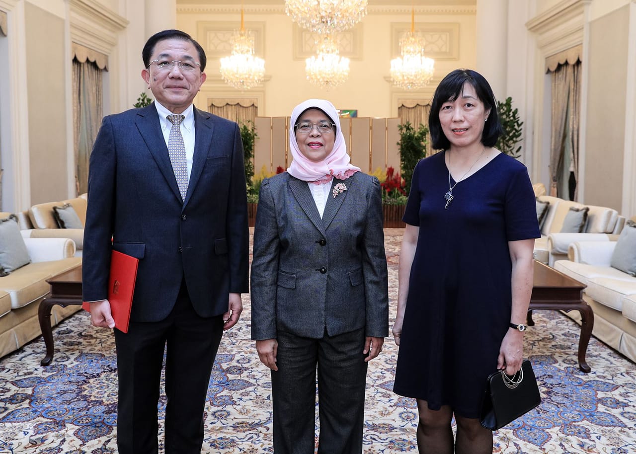 Three people stand in a room with chandeliers and sofas. Halimah Yacob is centered, wearing a headscarf and grey suit.