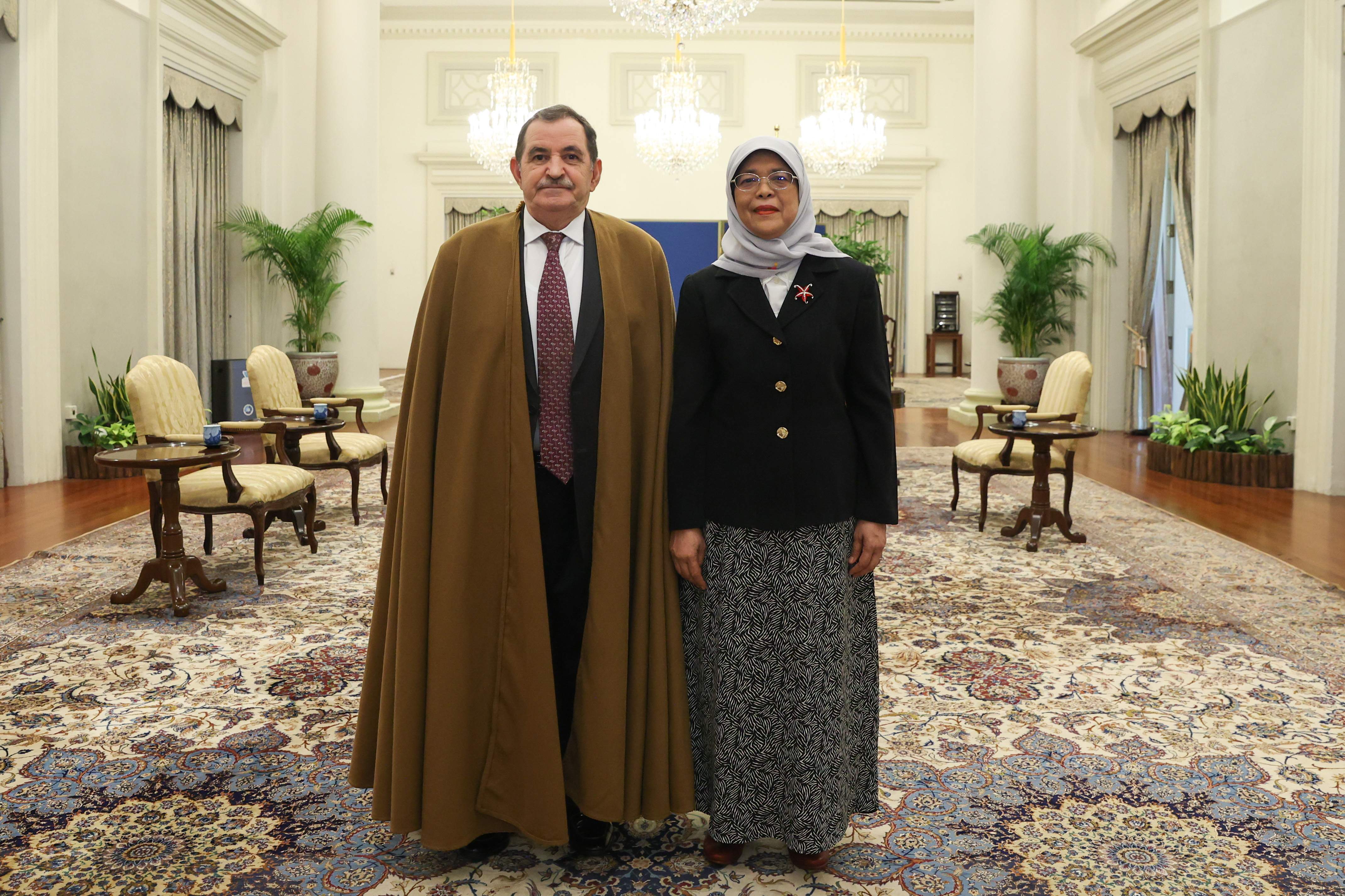 Two people standing in a room, man in brown robe, woman in hijab and black blazer.