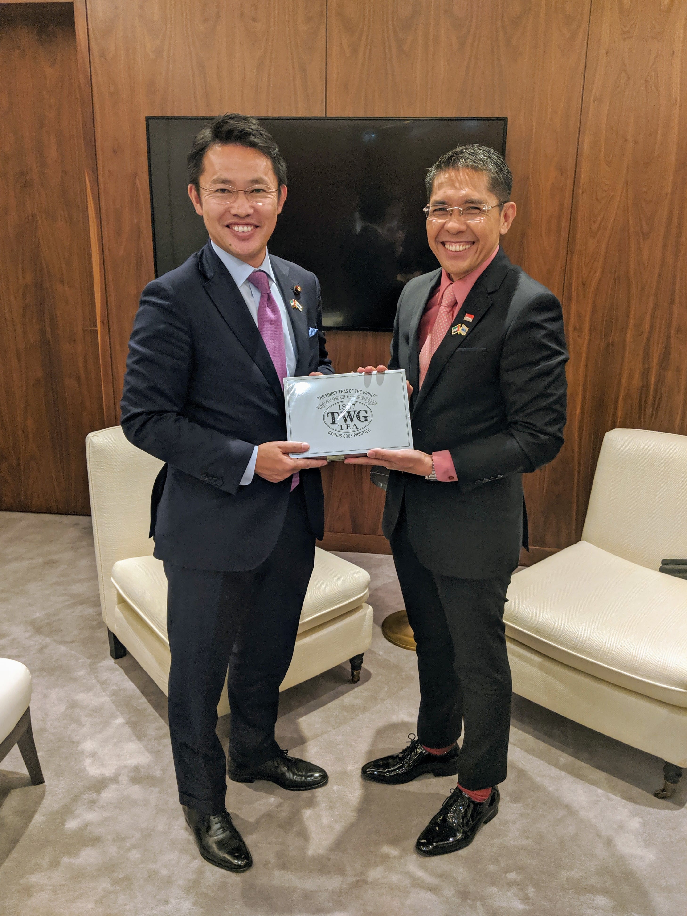 Two men in suits holding a TWG Tea box; flags on lapels, neutral colored furniture, wood wall.