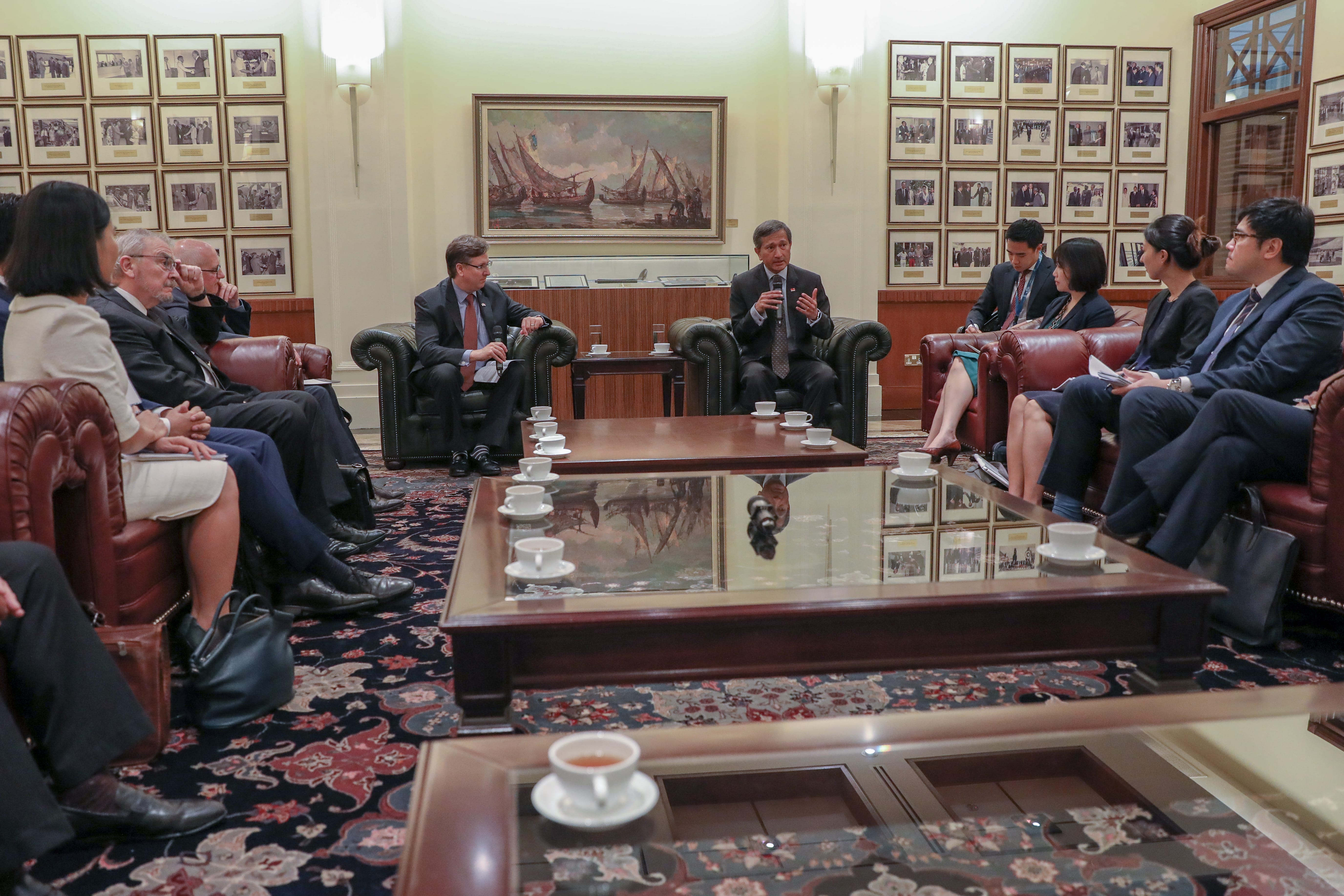 Meeting in a room with people seated on leather sofas, tea cups on a table, and framed pictures on the wall.