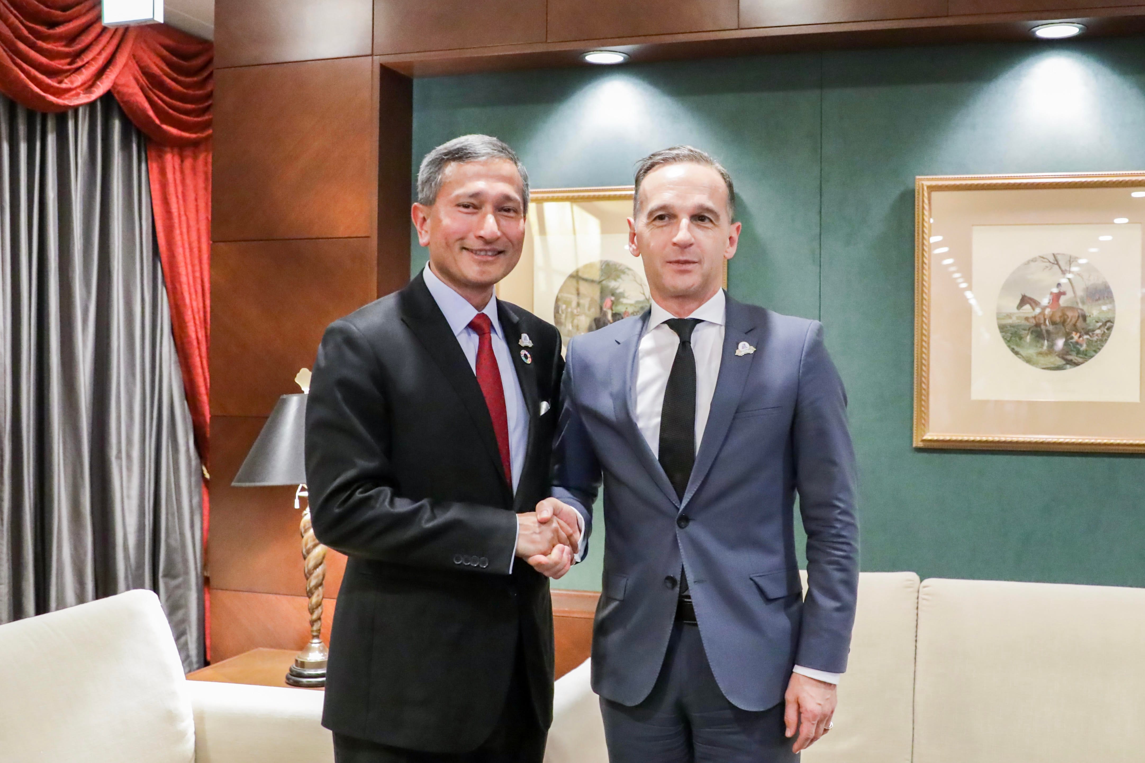 Two men in suits shaking hands, indoors. One in black with a red tie, the other in gray with a black tie.