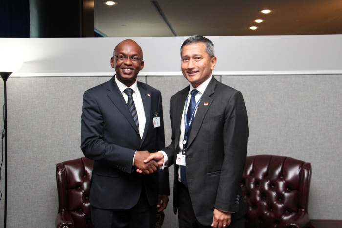 Two men in suits shake hands in front of gray wall with chairs.