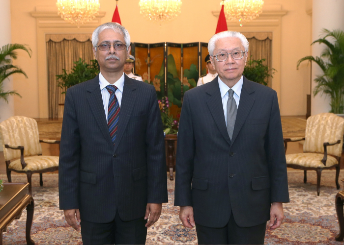 Two men in suits stand in a room with flags and a patterned screen.