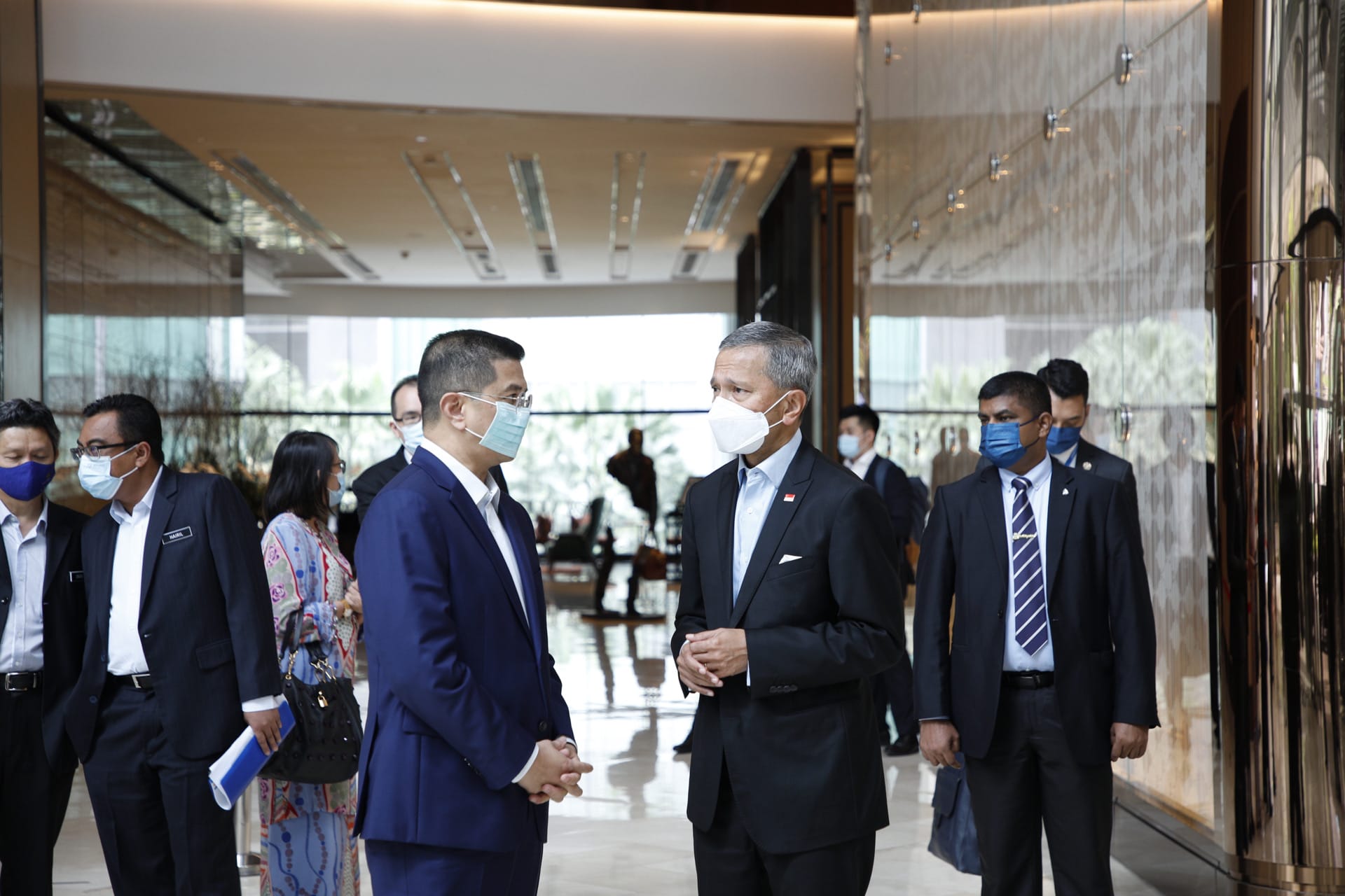 Two masked men in suits converse in a modern lobby with other people present.