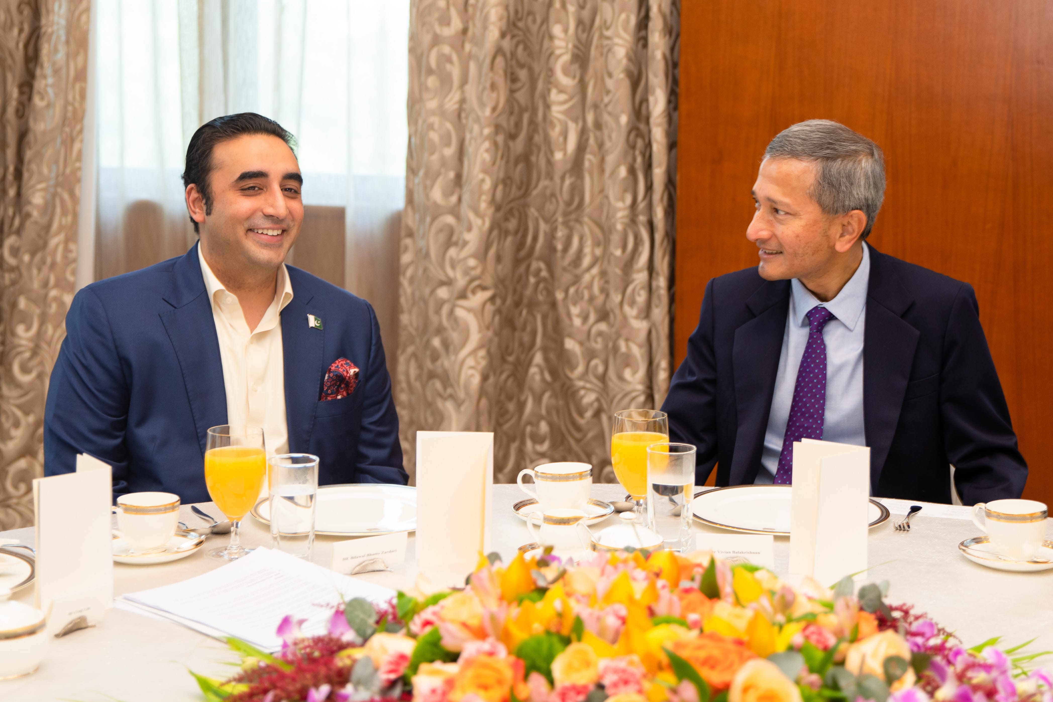 Two men in suits seated at a formal dining table with drinks, flowers, and place settings.