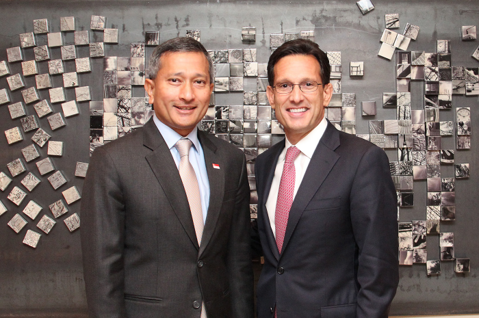 Two men in suits stand before a square-tiled wall; one wears a Singapore flag pin.