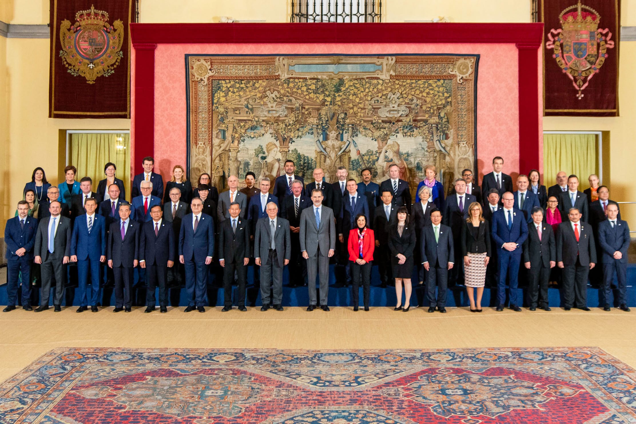Formal group portrait of suited men and women before tapestries in a palace setting.