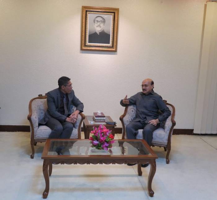 Two men in suits sit facing each other in ornate chairs, flower bouquet on table. Portrait hangs above.