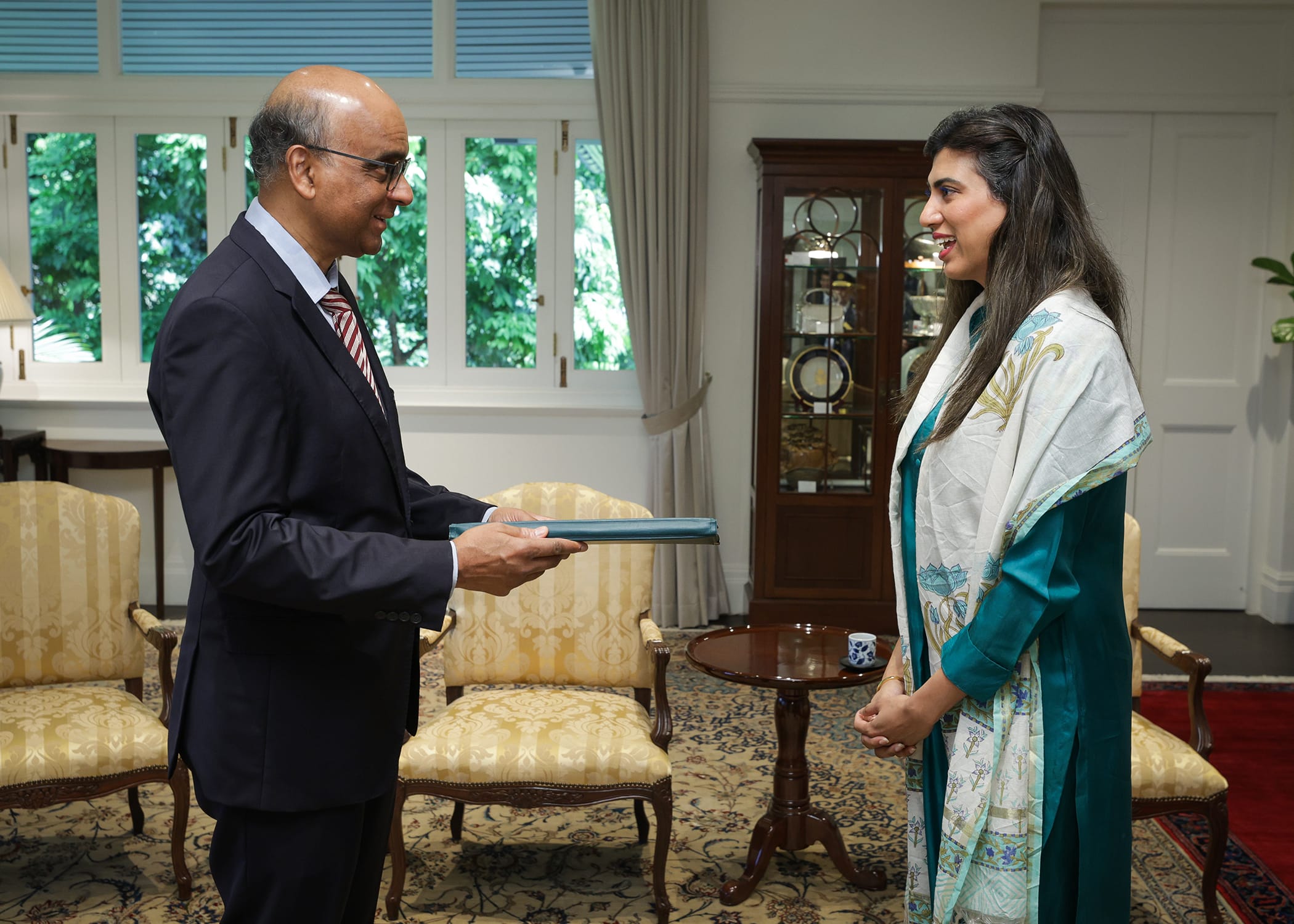 Man in suit presents document to woman in green outfit in ornate room.