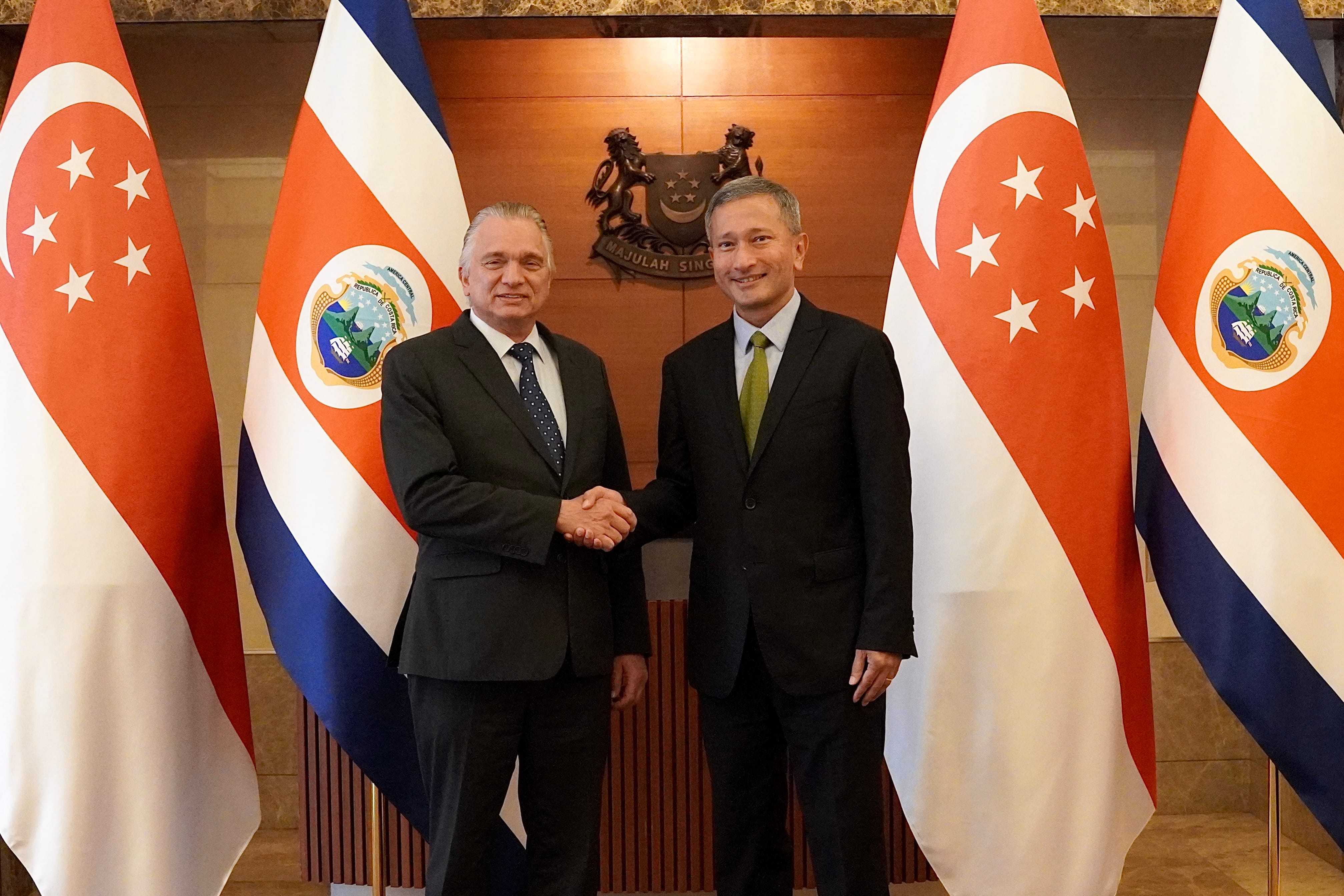 Two men in suits shake hands, between flags of Costa Rica and Singapore.