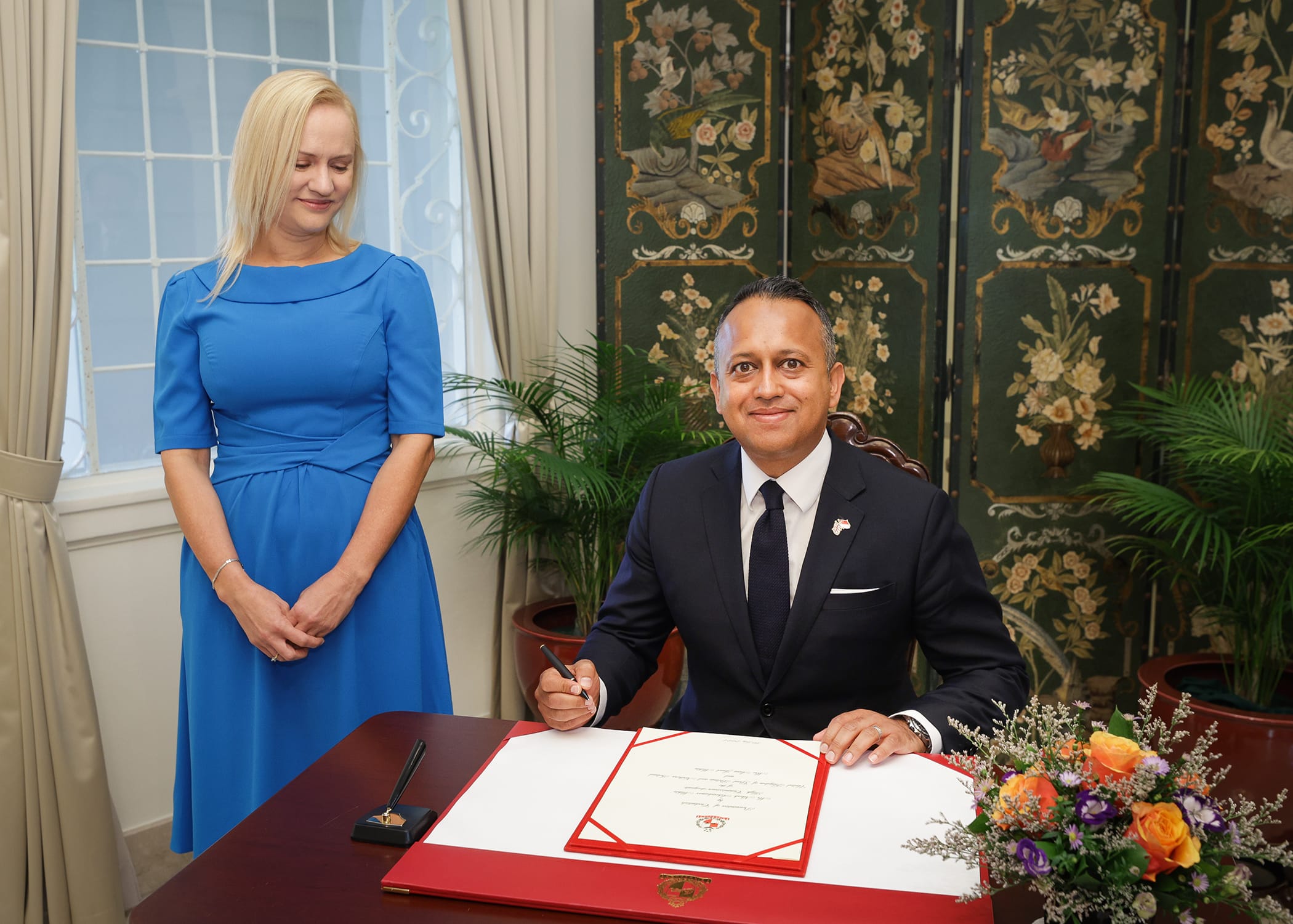 Man in suit signing document on desk, woman in blue dress stands nearby. Floral decorations present.