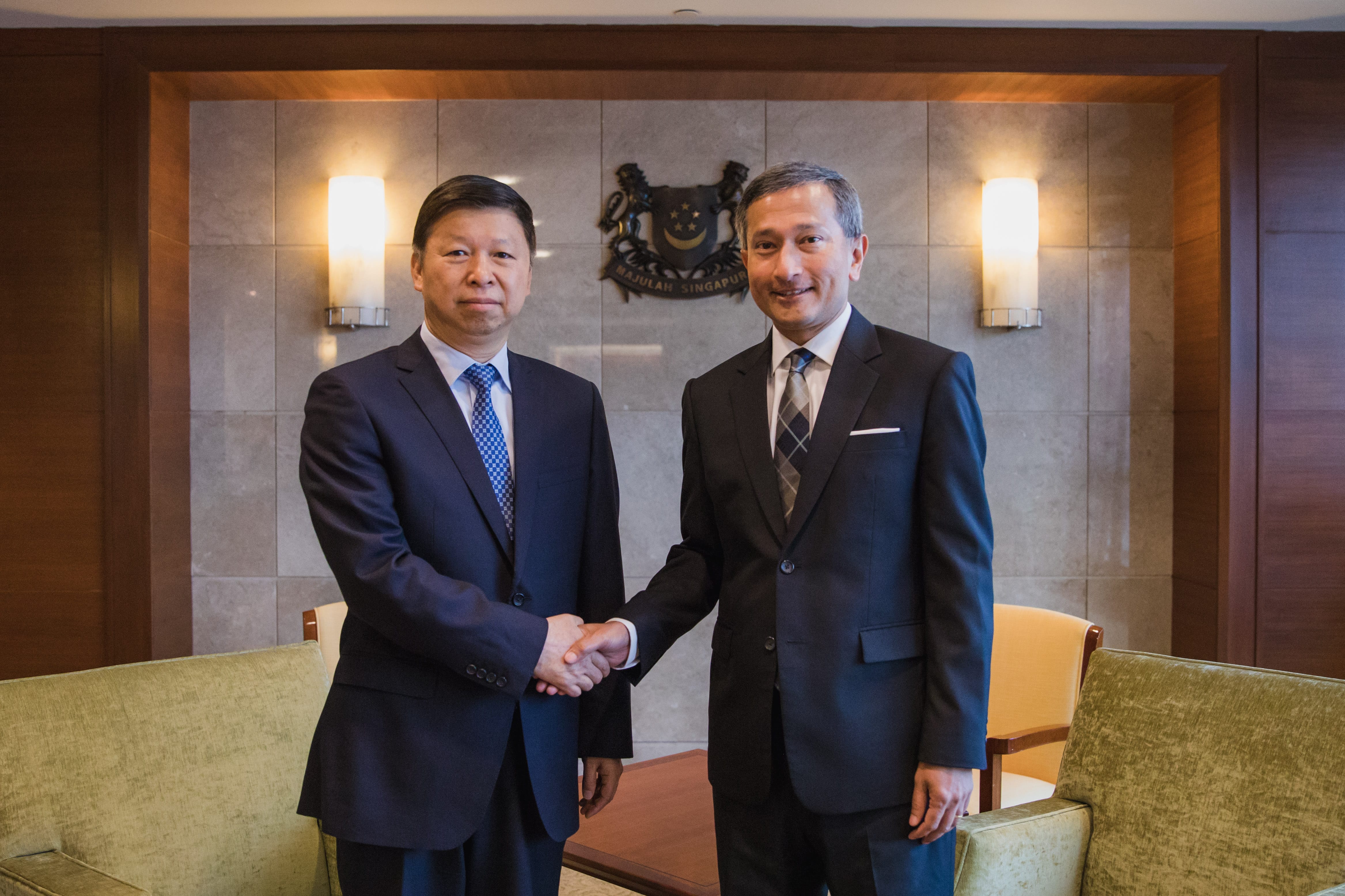 Two men in suits shaking hands, with the Singapore coat of arms visible in the background.