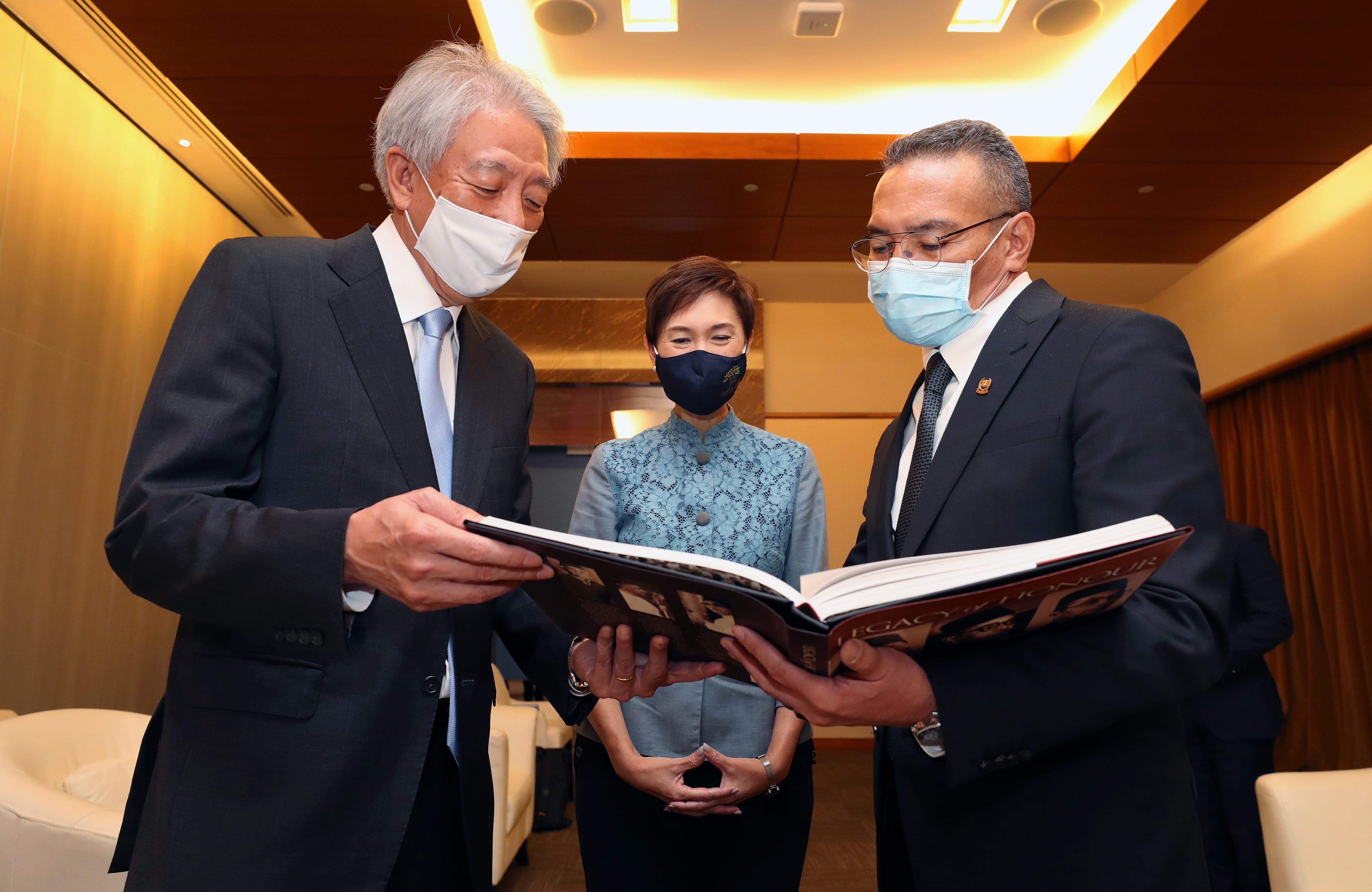 Three masked people examining an open book, "Legacy of Honour", in an indoor setting.