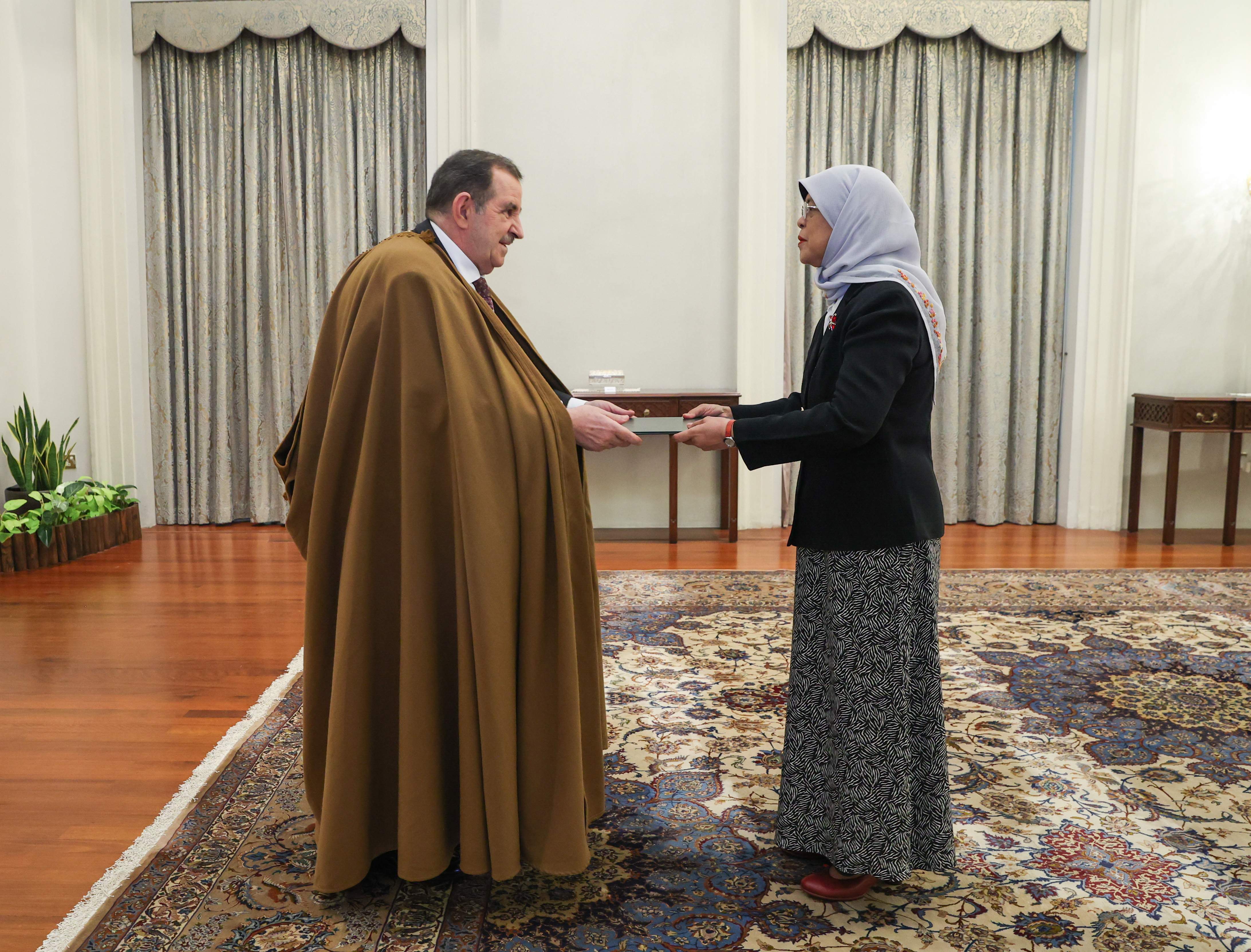 Two people exchanging a small, rectangular object, indoors on an ornate carpet.