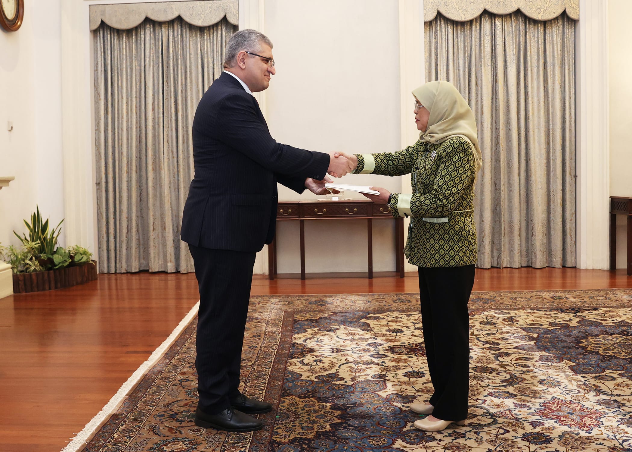 Two people shaking hands in a formal room, one holding papers.