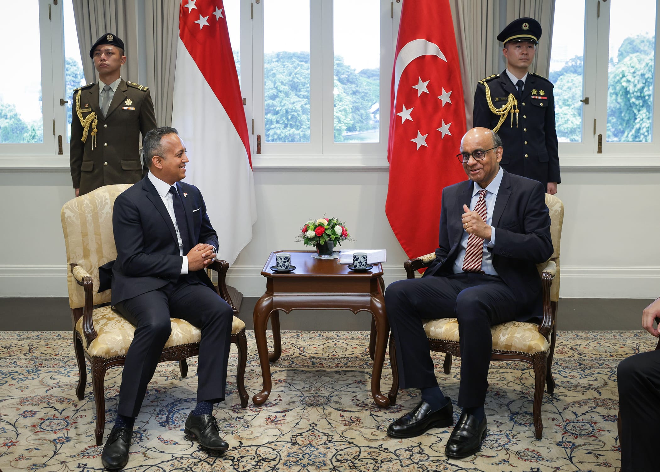 Two seated men in suits, flanked by two standing men in uniform, Singapore flags, ornate rug.