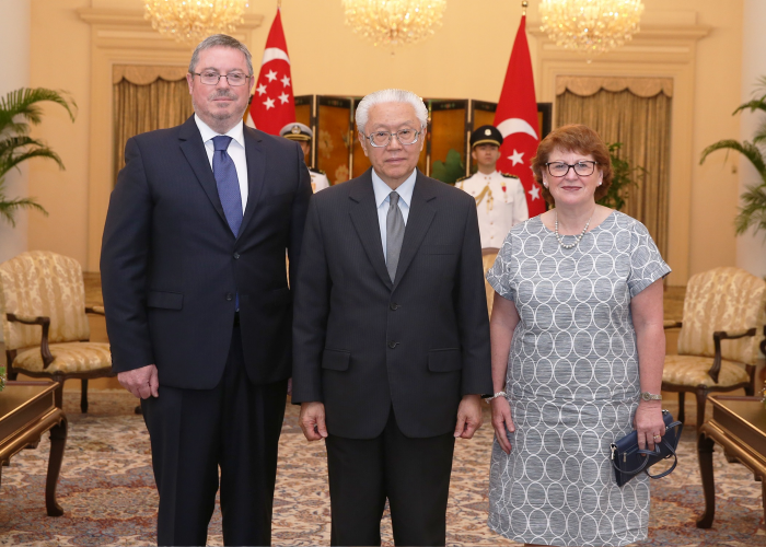 Three people pose indoors with Singapore flags; two in suits, one in patterned dress.