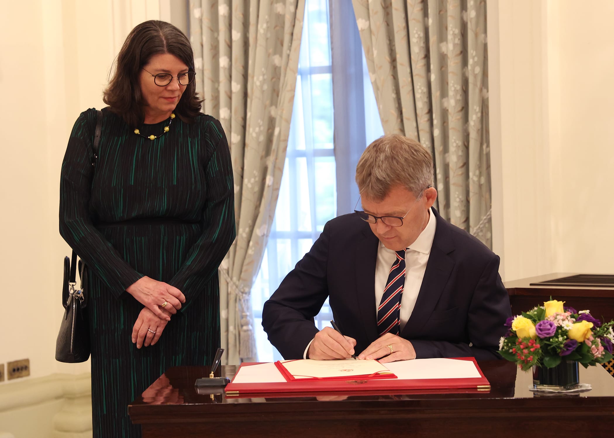 Person signing document on desk as woman watches. Floral arrangement.