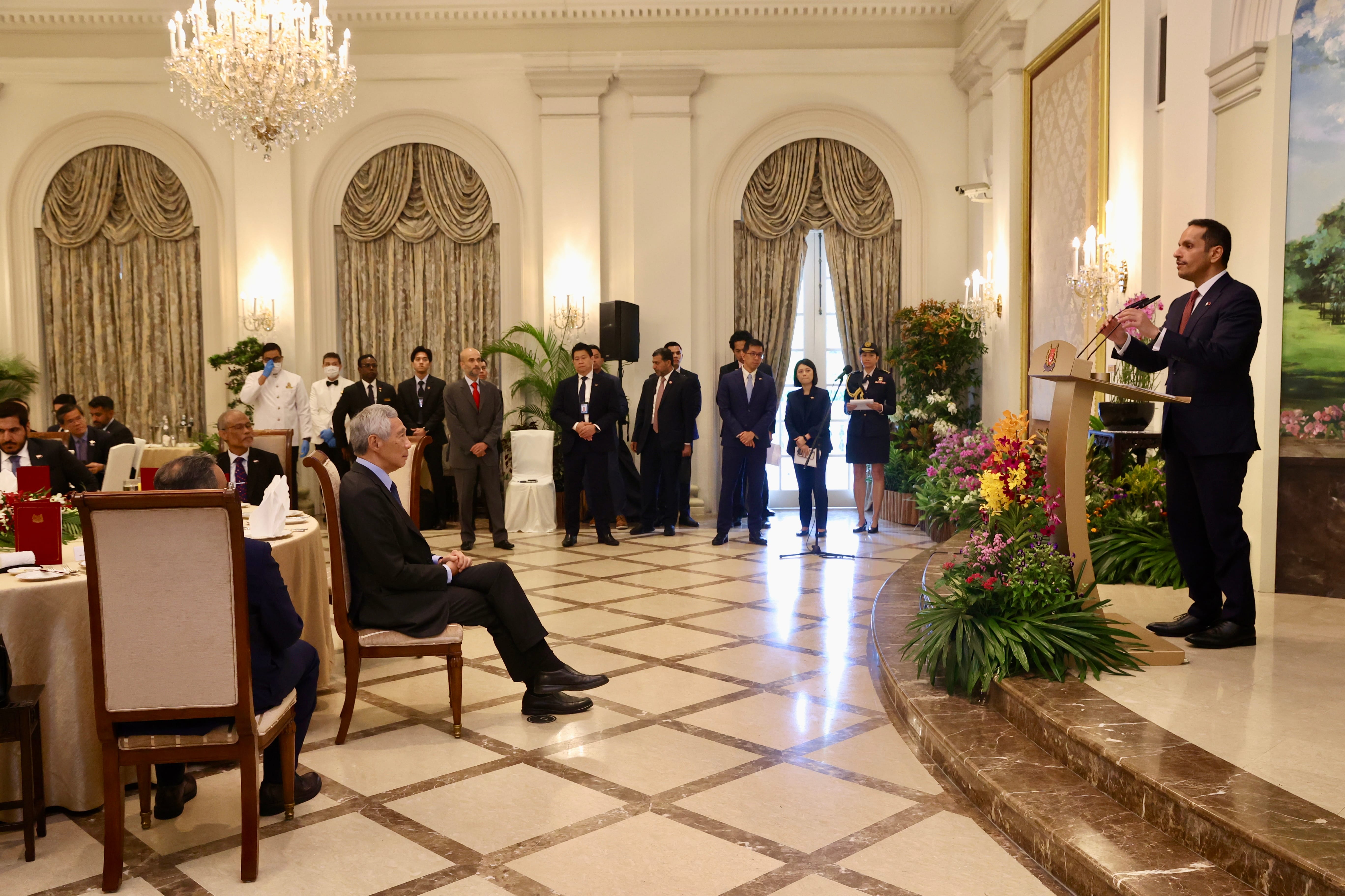 Man at lectern addresses room. Attendees seated and standing. Chandelier overhead.