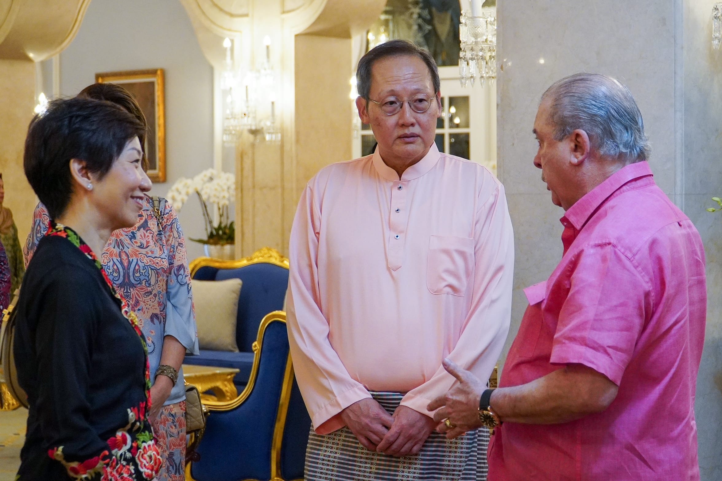 Three people in an ornate room; one in pink shirt, glasses, striped sarong.